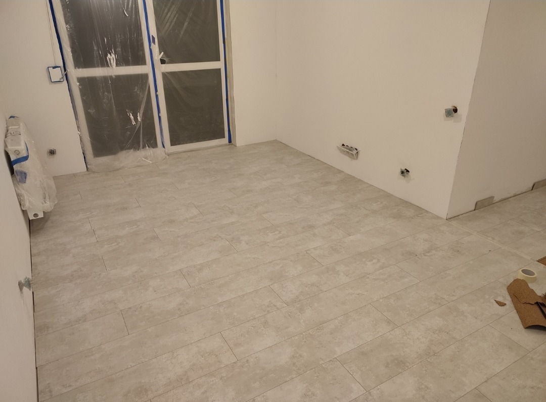 Wykończona podłoga z płytek imitujących drewno w jasnym odcieniu, widoczne gniazdka elektryczne w ścianie, okno zabezpieczone folią malarską, pozostałości po pracach wykończeniowych w rogu...