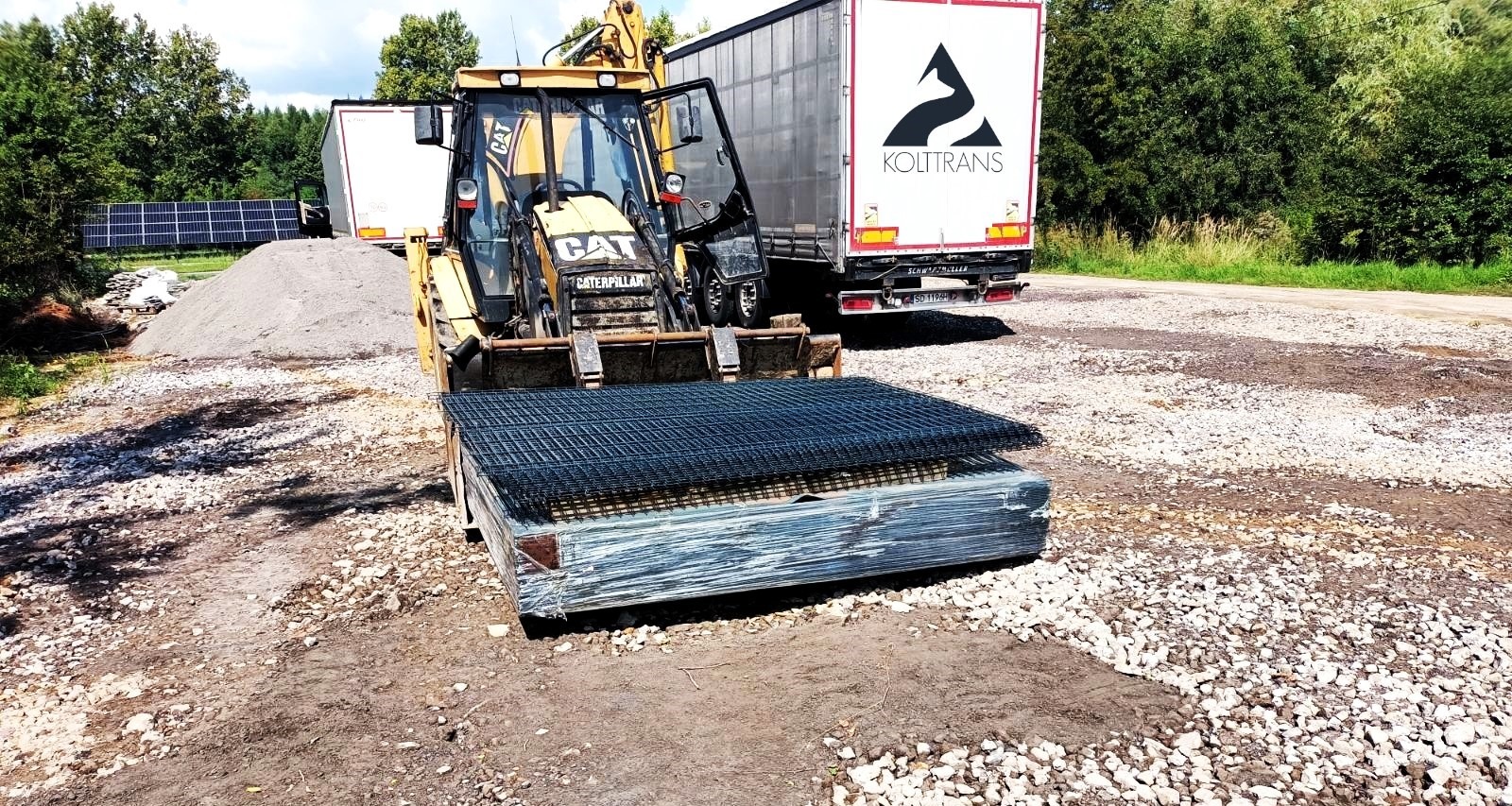 Żółta ładowarka CAT przewozi paletę z siatką zbrojeniową na żwirowym placu budowy, w tle zaparkowana ciężarówka Koultrans i panele słoneczne.
