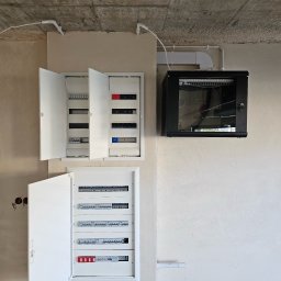 CleverHouse Paweł Piotrowski - Rozdzielnice elektryczne (otwarte) i szafa rack na ścianie w surowym wnętrzu. Widoczne kable i przewody. Instalacja w trakcie realizacji.