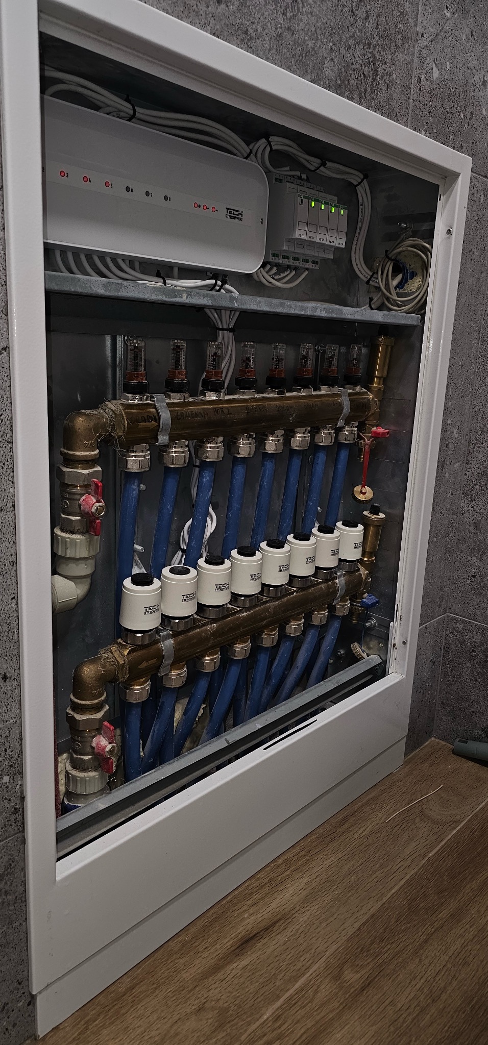 Rozdzielacz ogrzewania podłogowego w szafce, z widocznymi rurami, zaworami termostatycznymi i modułem sterującym TECH, zamontowany na ścianie z szarych płytek.