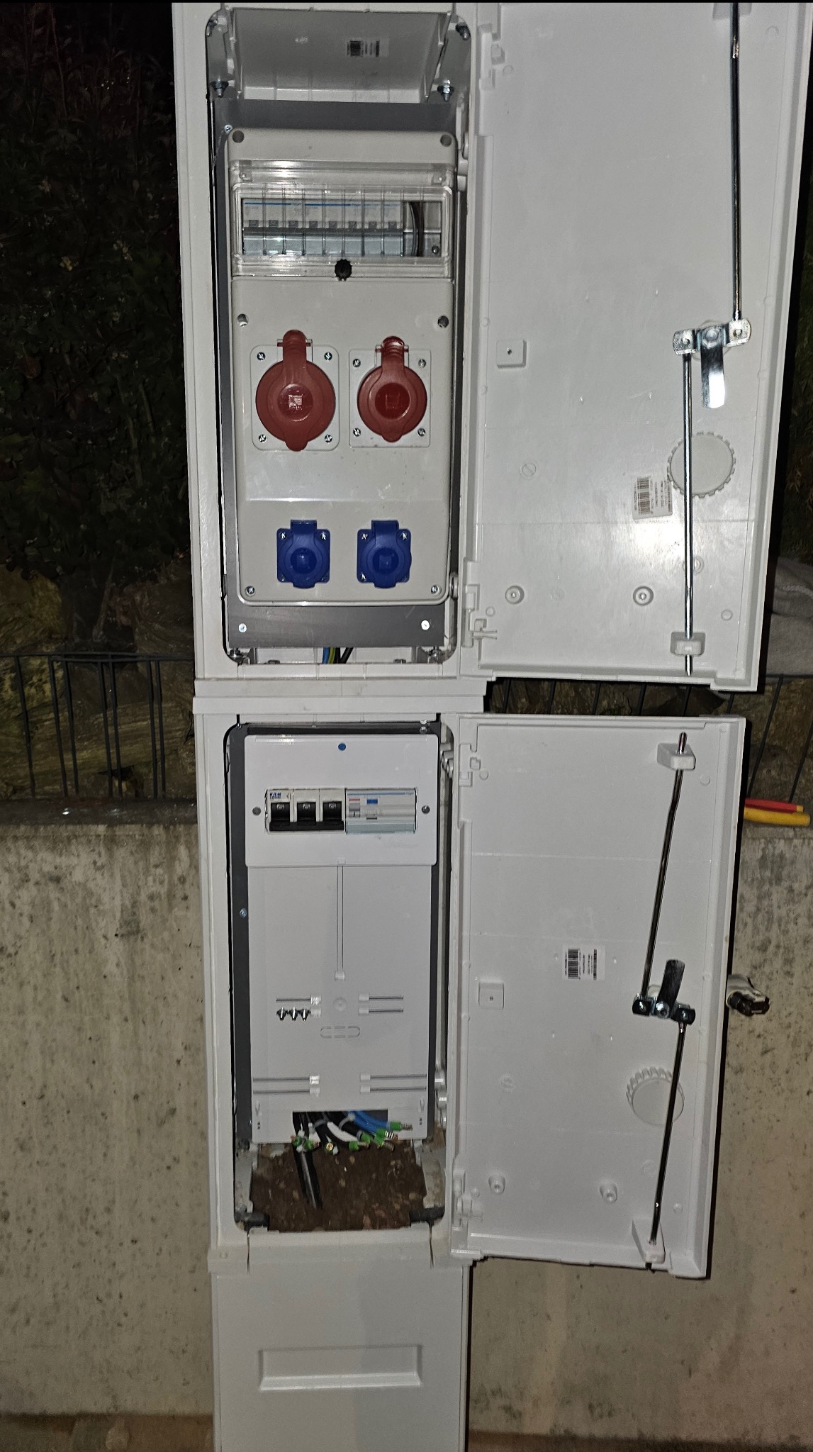 Otwarta, dwuczęściowa skrzynka elektryczna z gniazdami (czerwone i niebieskie) i bezpiecznikami, zamontowana na zewnątrz, z widocznymi kablami w dolnej części.