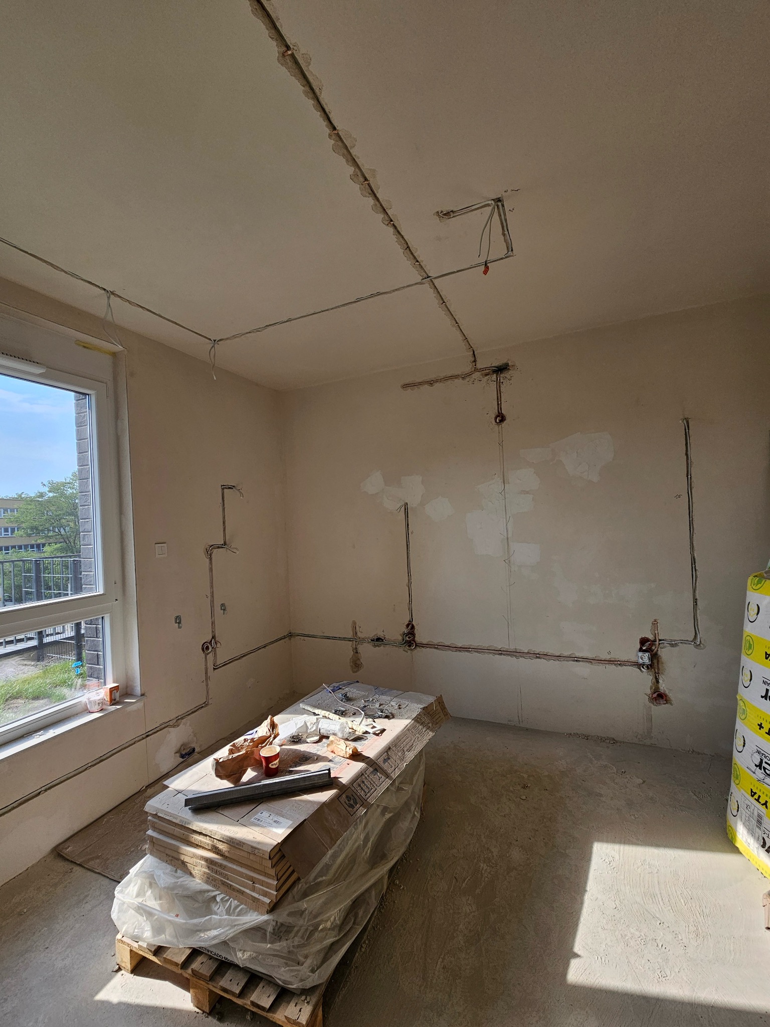 Wnętrze w trakcie prac elektrycznych: kable rozprowadzone po ścianach i suficie, przygotowane punkty instalacyjne. Częściowo wykończone ściany, okno z widokiem na zewnątrz.