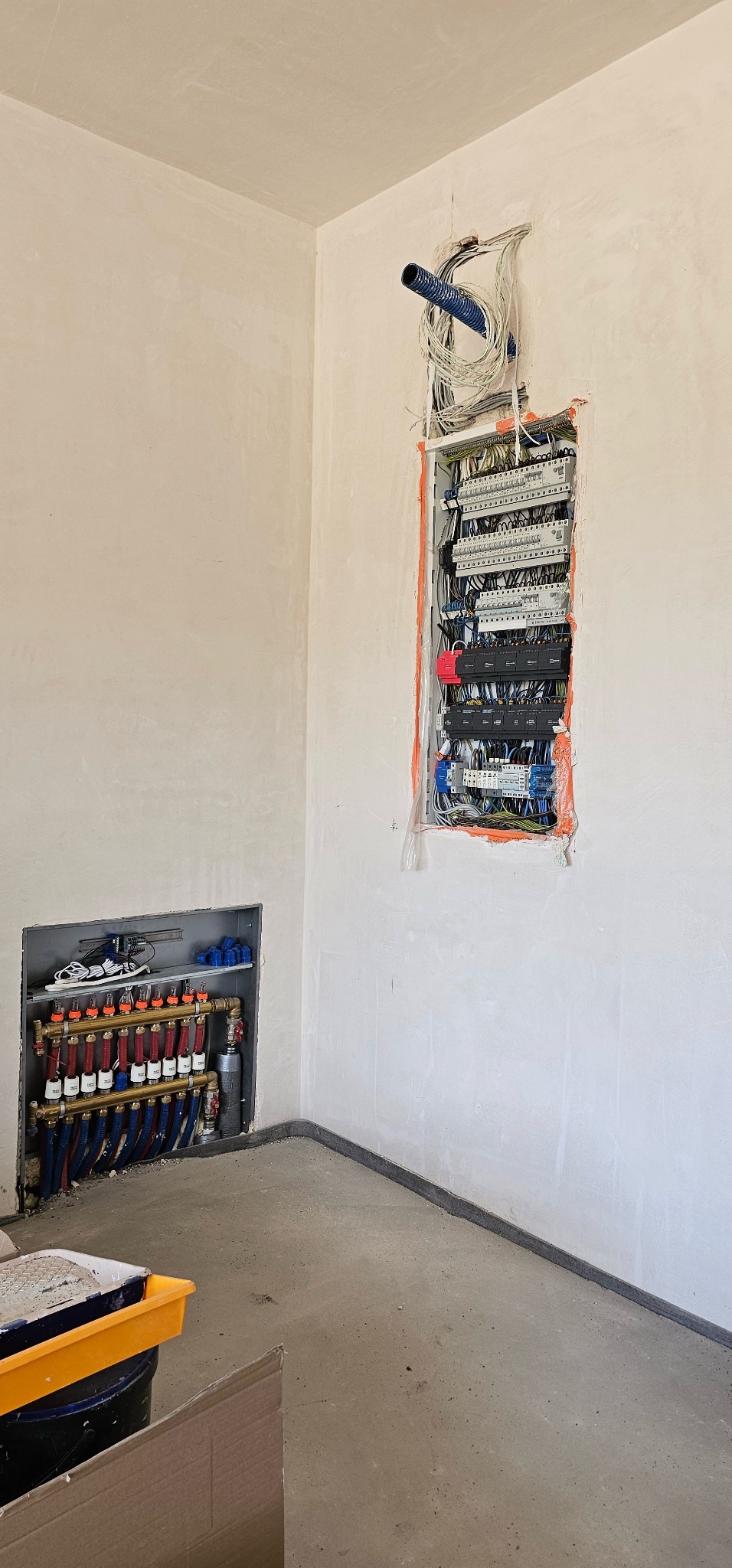 Rozdzielnia elektryczna z przewodami w ścianie i instalacja ogrzewania podłogowego w surowym pomieszczeniu, widok z dołu, w tle ściany w trakcie wykończenia.