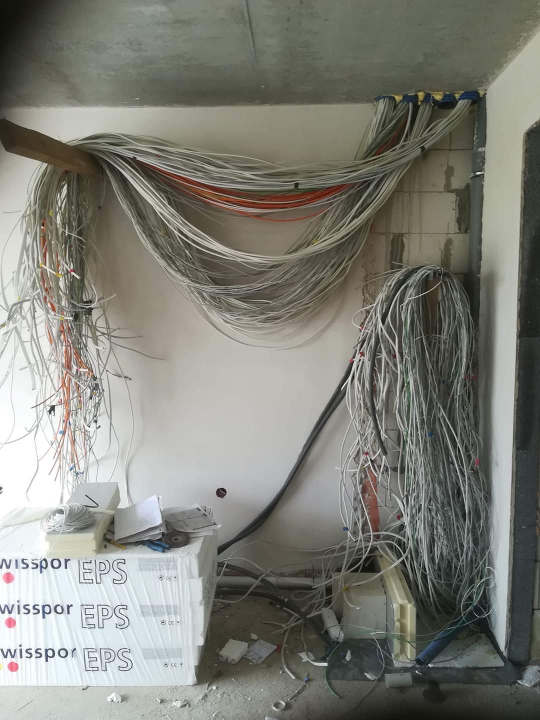 Rozciągnięte kable elektryczne w surowym wnętrzu, częściowo podwieszone na drewnianej belce, z widocznymi puszkami instalacyjnymi i materiałami izolacyjnymi.