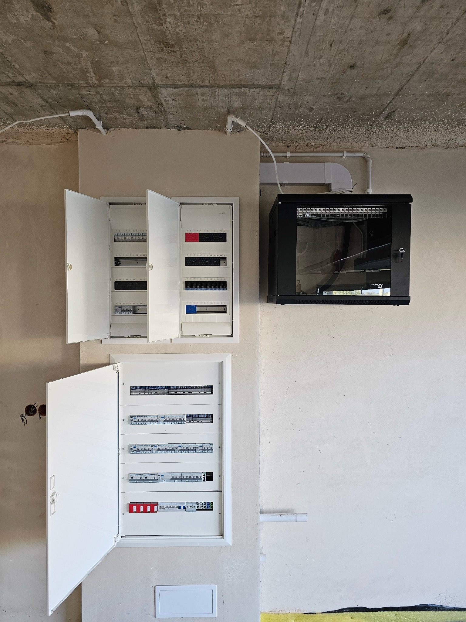 Rozdzielnice elektryczne (otwarte) i szafa rack na ścianie w surowym wnętrzu. Widoczne kable i przewody. Instalacja w trakcie realizacji.