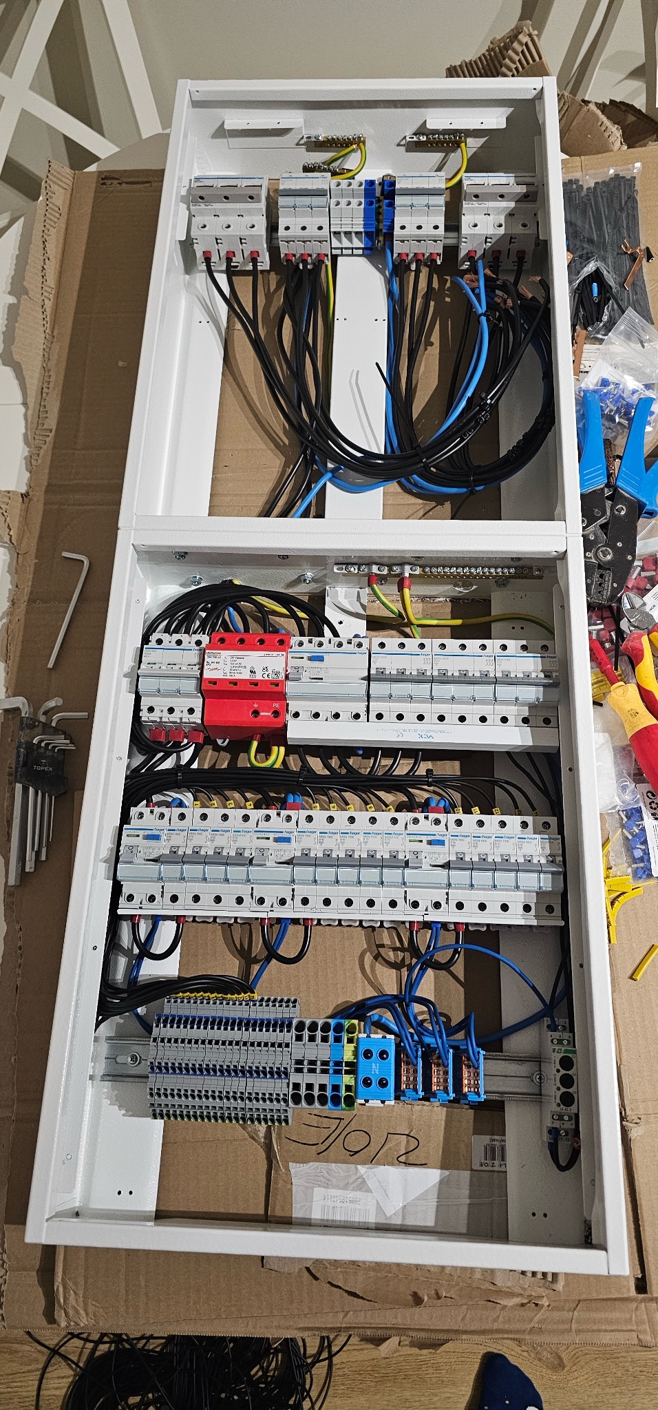 Rozdzielnica elektryczna w białej obudowie, widoczne zabezpieczenia, przewody i listwy zaciskowe. Częściowo widoczne narzędzia monterskie.