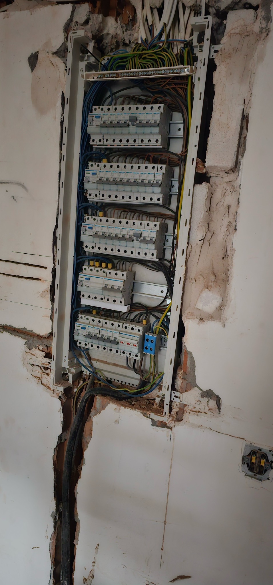 Rozdzielnica elektryczna w trakcie montażu w ścianie, widoczne kable i zabezpieczenia. Ściana w stanie surowym, z widocznymi śladami po kuciu.