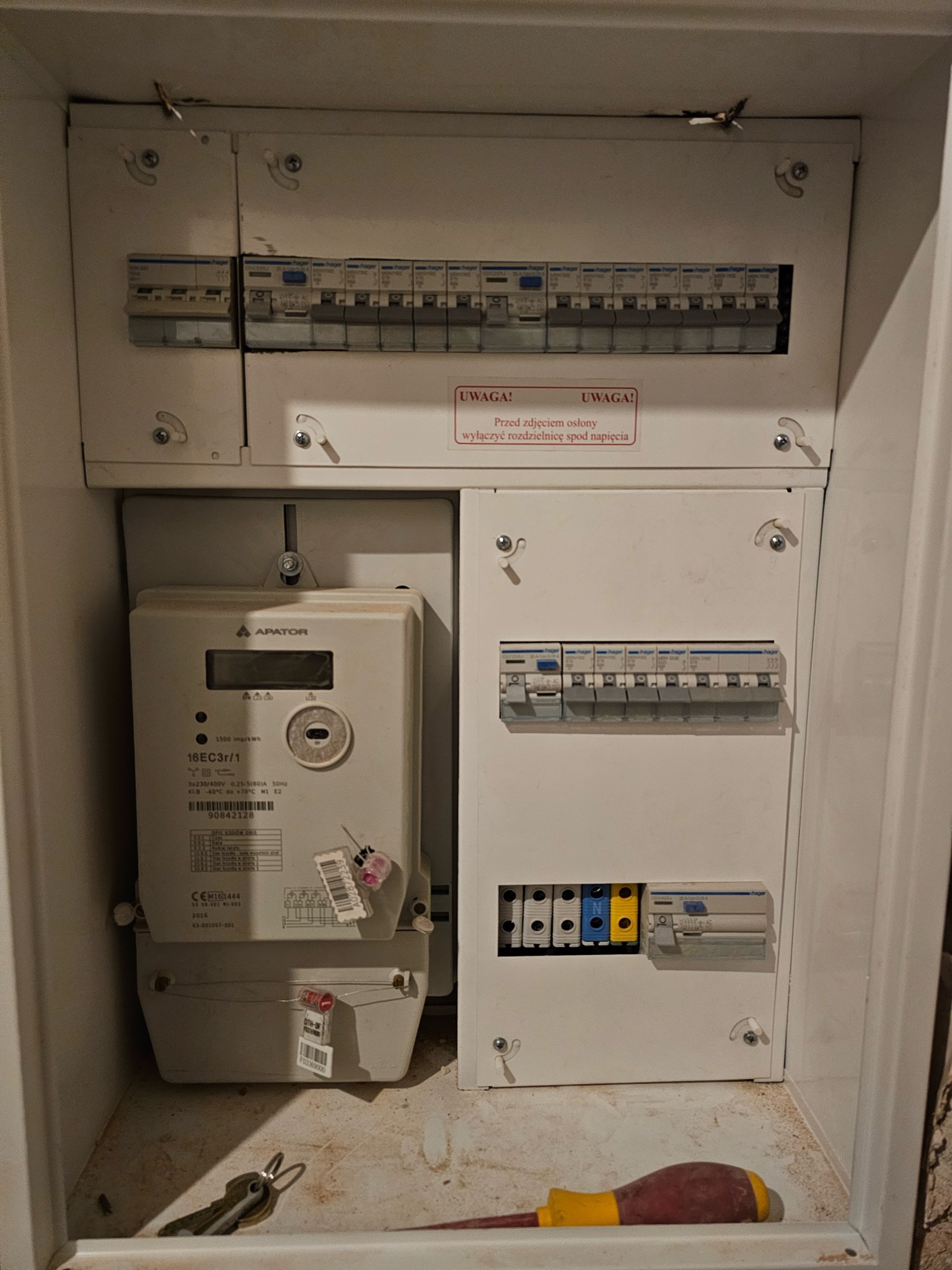 Rozdzielnia elektryczna z licznikiem Apator, bezpiecznikami Hager i żółto-czerwonym śrubokrętem. Widoczne ostrzeżenia o wyłączeniu napięcia przed zdjęciem osłony.