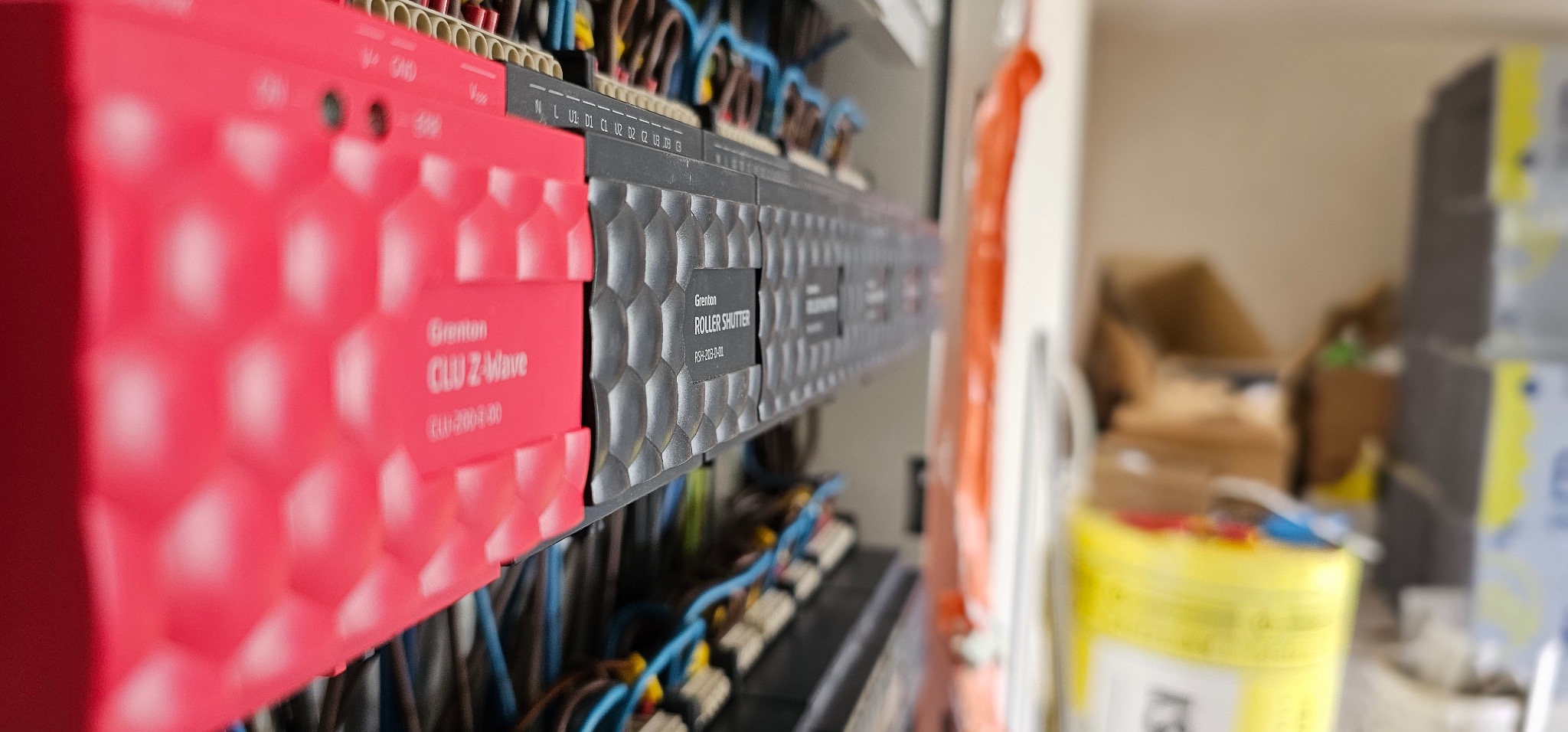 Panel sterowania inteligentnego domu Grenton z modułami CLU Z-Wave i ROLLER SHUTTER, widoczne przewody i elementy instalacji, w tle kartony i wiadro.