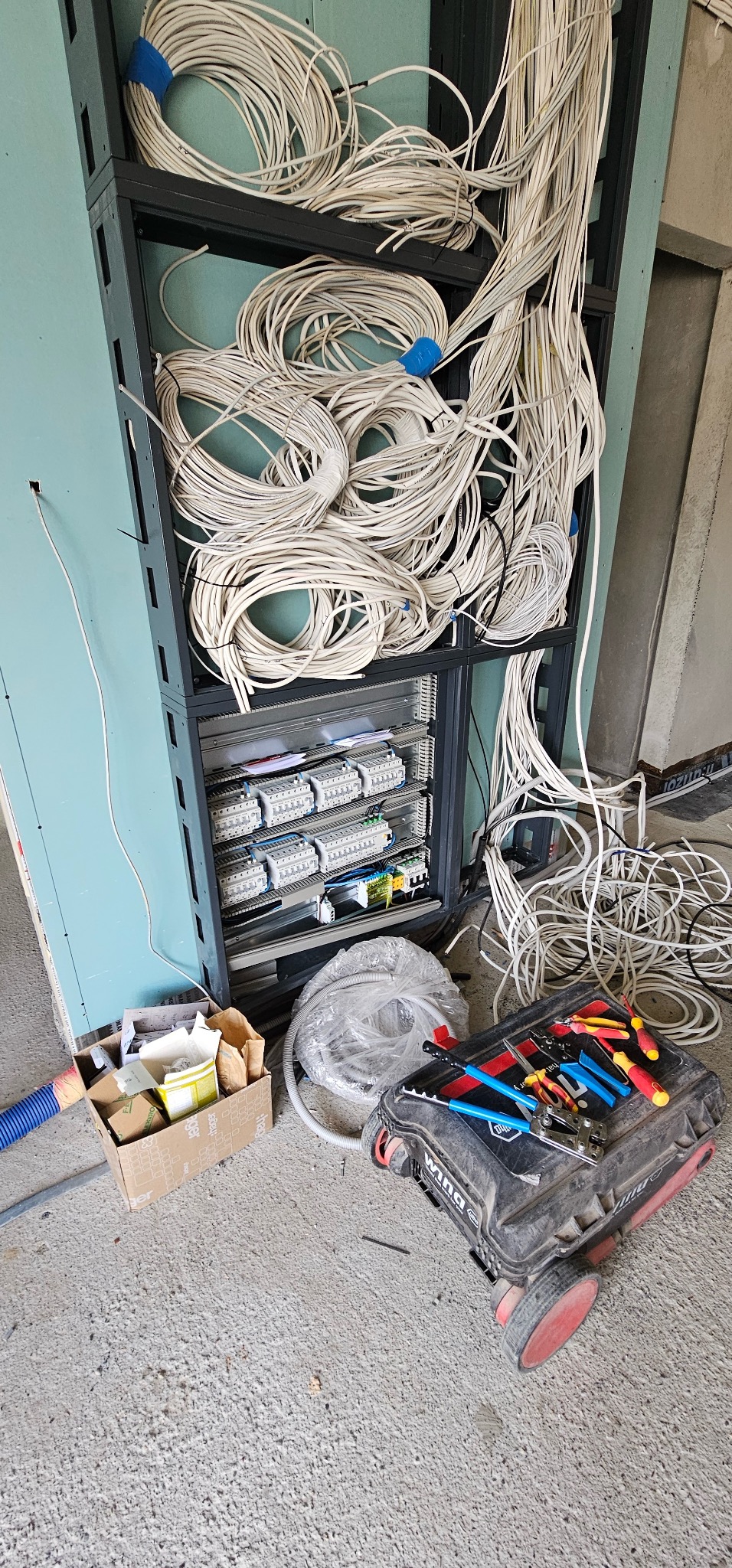 Rozdzielnia elektryczna w trakcie montażu, widoczne kable, skrzynka z narzędziami i elementy instalacji. Przewody ułożone w pętle, częściowo poza szafą.