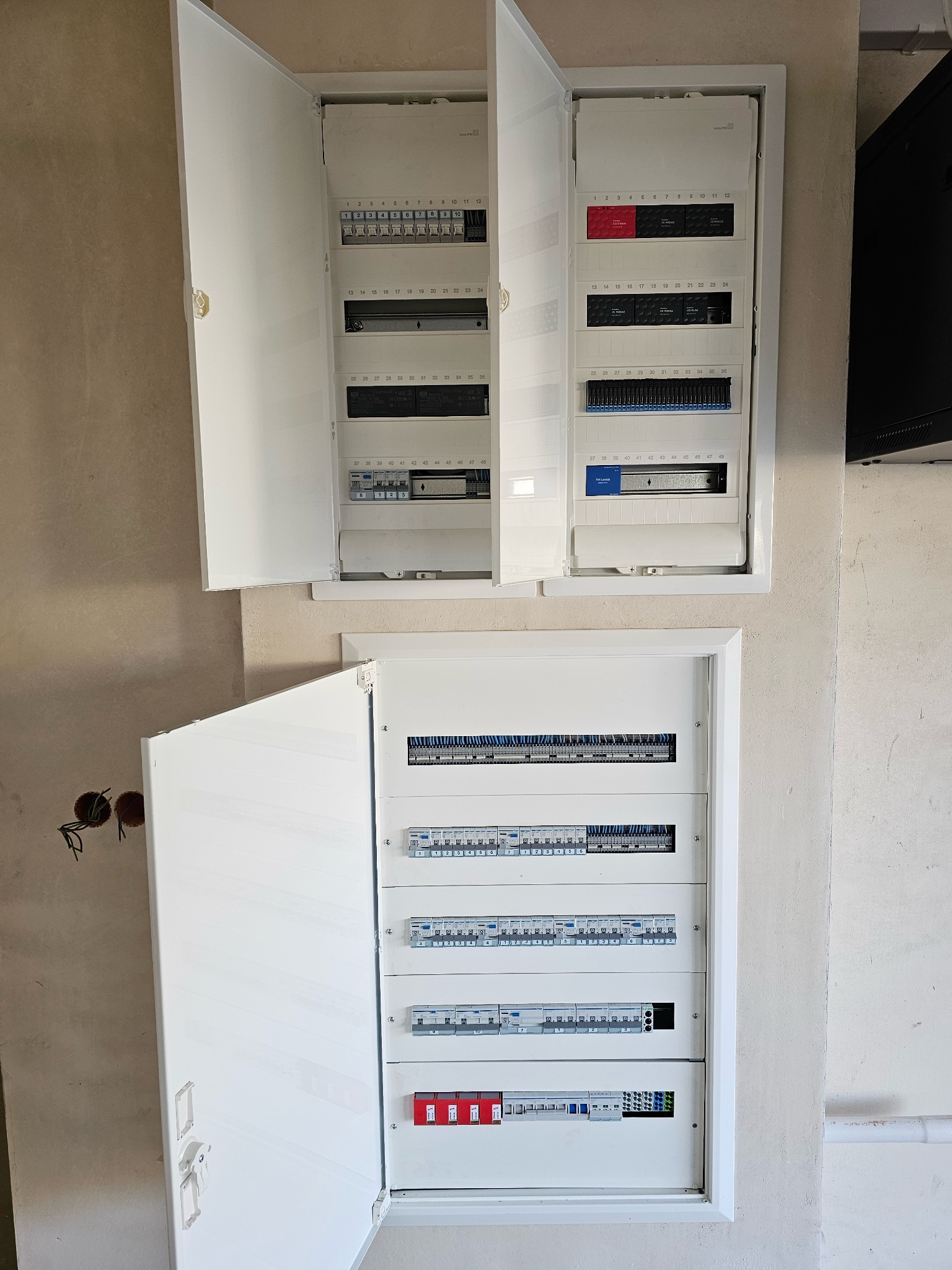 Dwa białe panele elektryczne z otwartymi drzwiczkami, widoczne zabezpieczenia i okablowanie. Jeden panel nad drugim, na surowej ścianie. Widoczne otwory na kable.