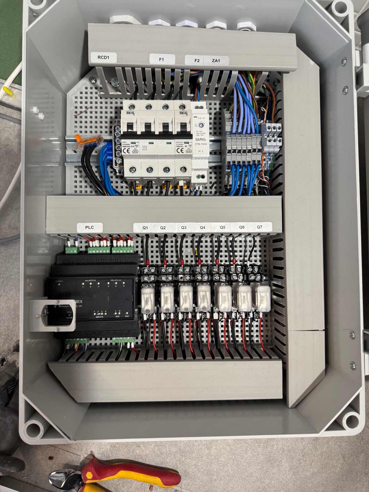 Szara skrzynka elektryczna z bezpiecznikami, przekaźnikami i przewodami. Widoczne oznaczenia RCD1, F1, F2, ZA1, PLC, Q1-Q7. U dołu obcięte narzędzia.