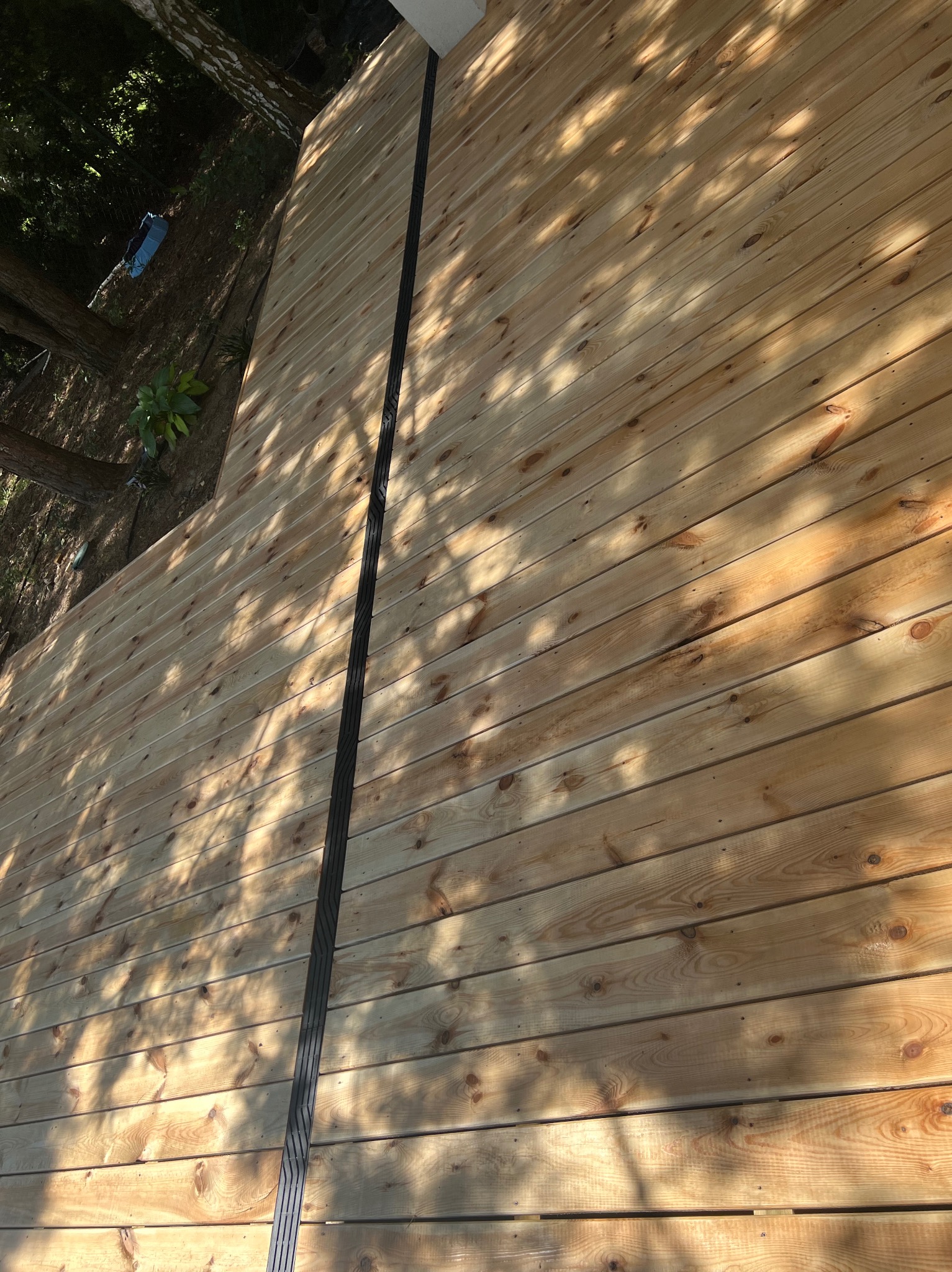 Świeżo ułożona deska tarasowa z widocznym profilem dylatacyjnym, oświetlona promieniami słonecznymi przebijającymi się przez drzewa.