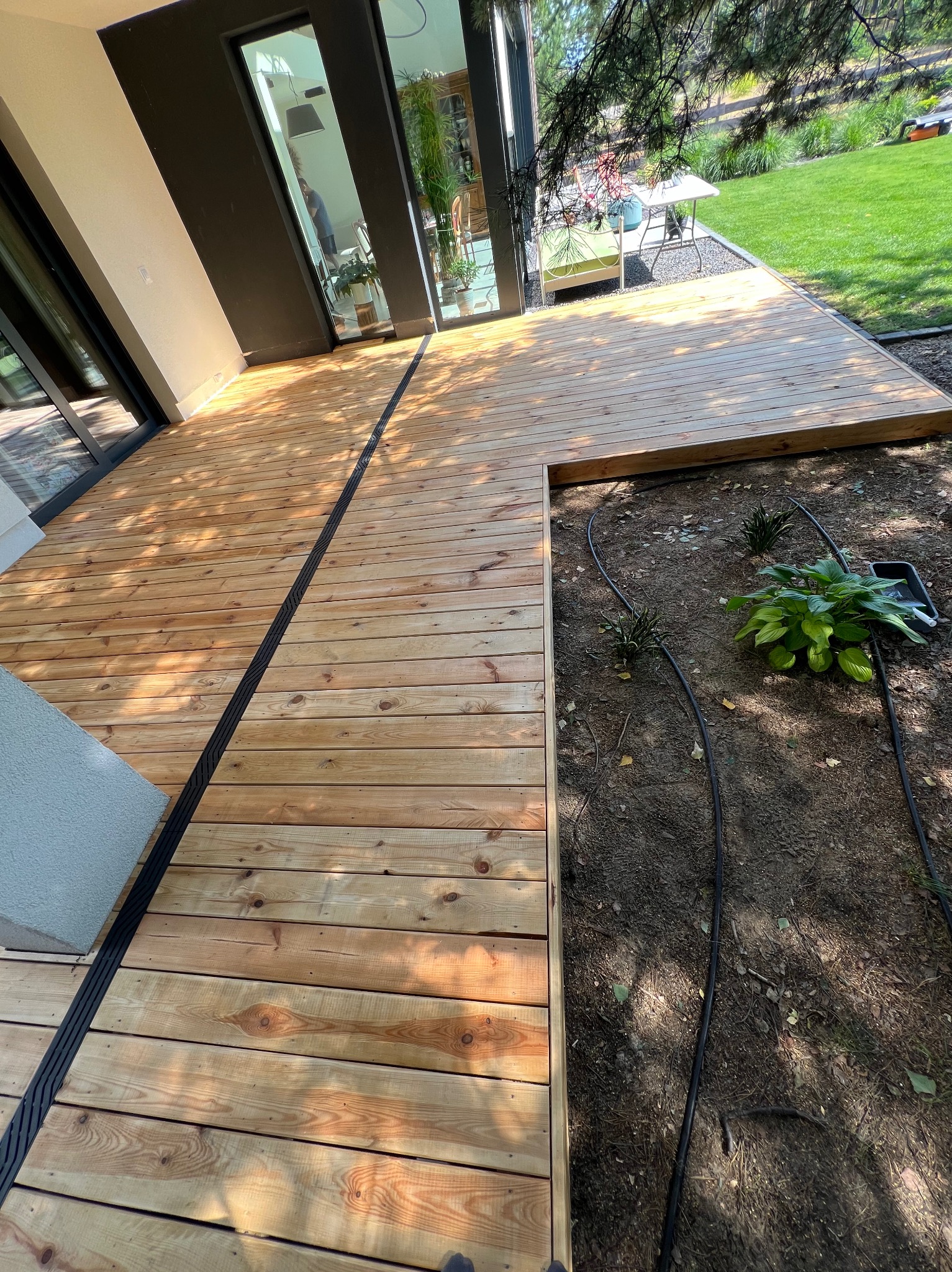 Nowoczesny taras drewniany z odpływem liniowym, przylegający do budynku z dużymi oknami, z widocznymi elementami nawadniania ogrodu.