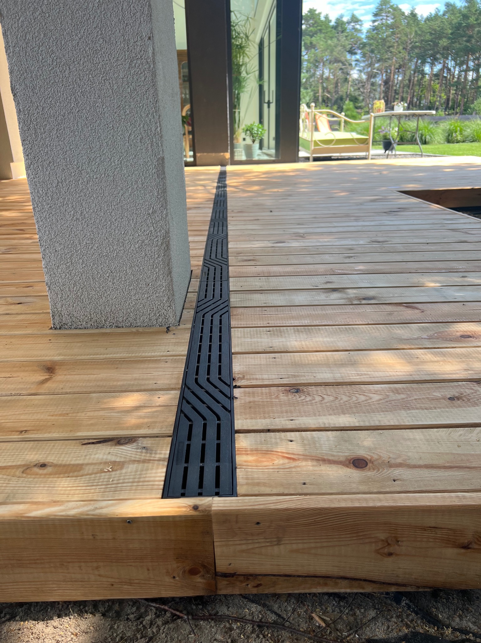 Nowoczesny taras z jasnego drewna z wbudowanym liniowym odpływem, widok na otaczający ogród i elementy wyposażenia.