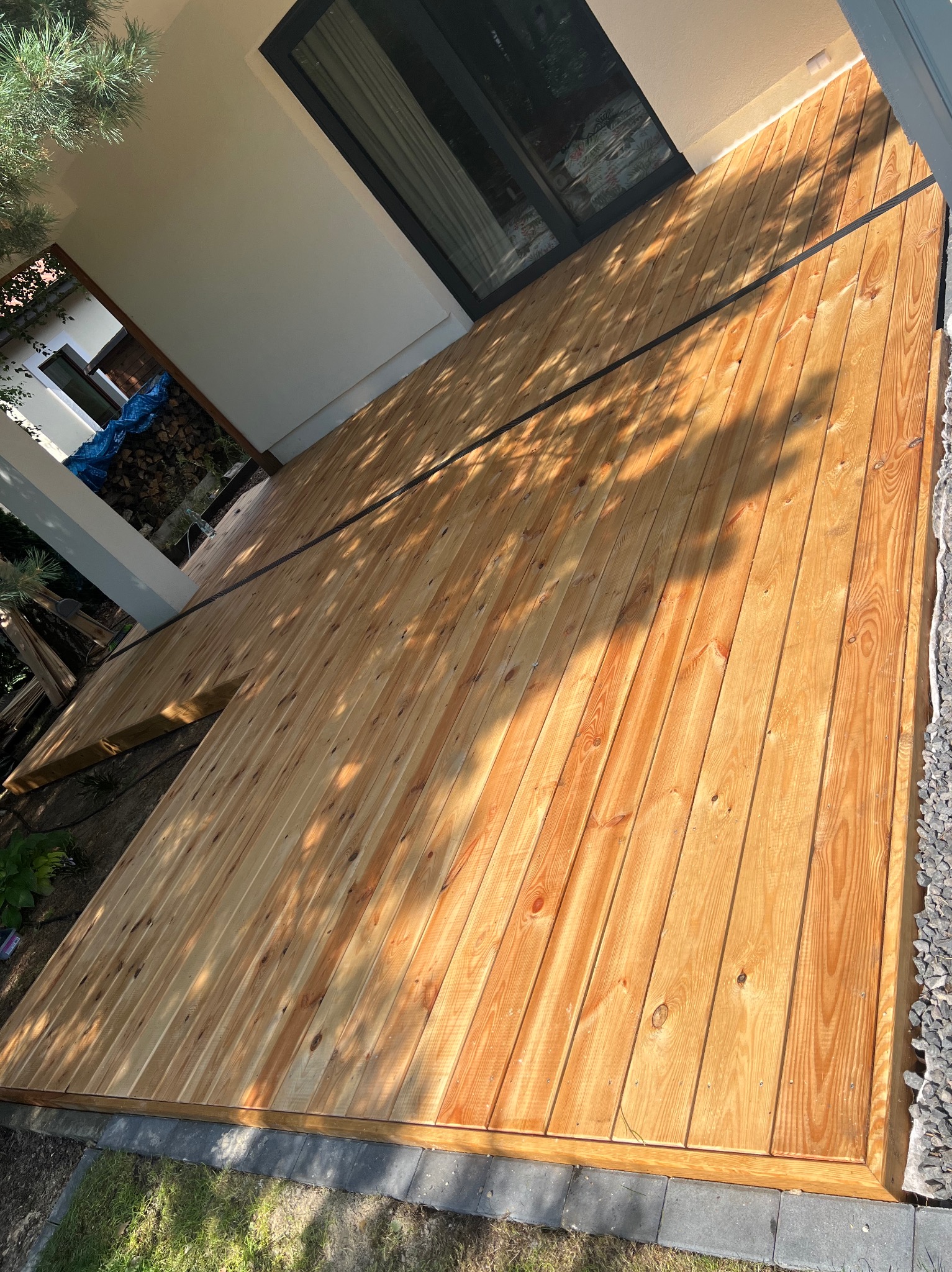 Nowo wybudowany taras z jasnego drewna, z widocznymi cieniami drzew, obrzeżami z szarego kamienia i fragmentem elewacji budynku z czarną ramą okienną.