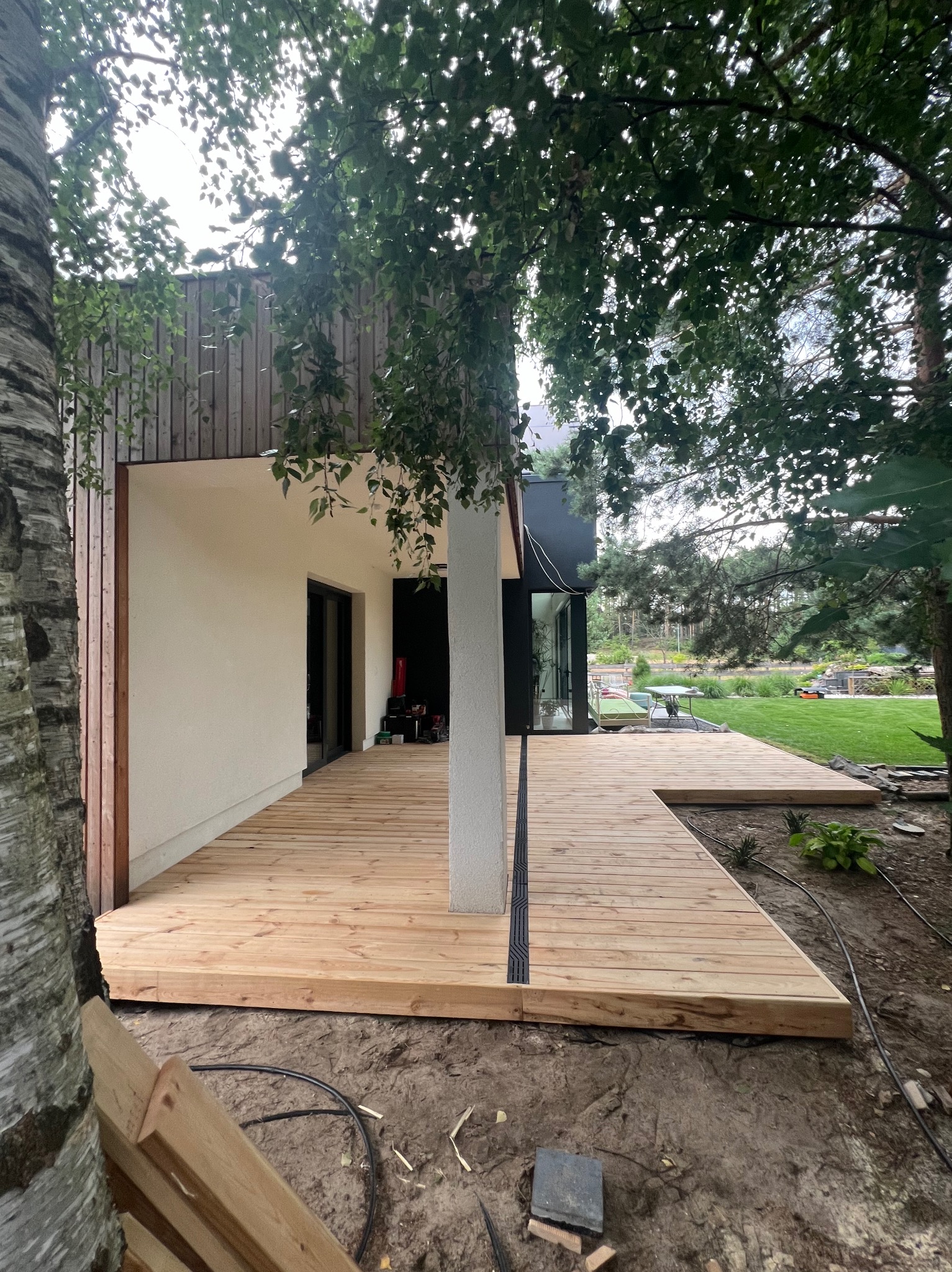 Nowoczesny dom z jasną, drewnianą konstrukcją tarasu, częściowo otoczonego ziemią i drzewami, z widocznym systemem odprowadzania wody.