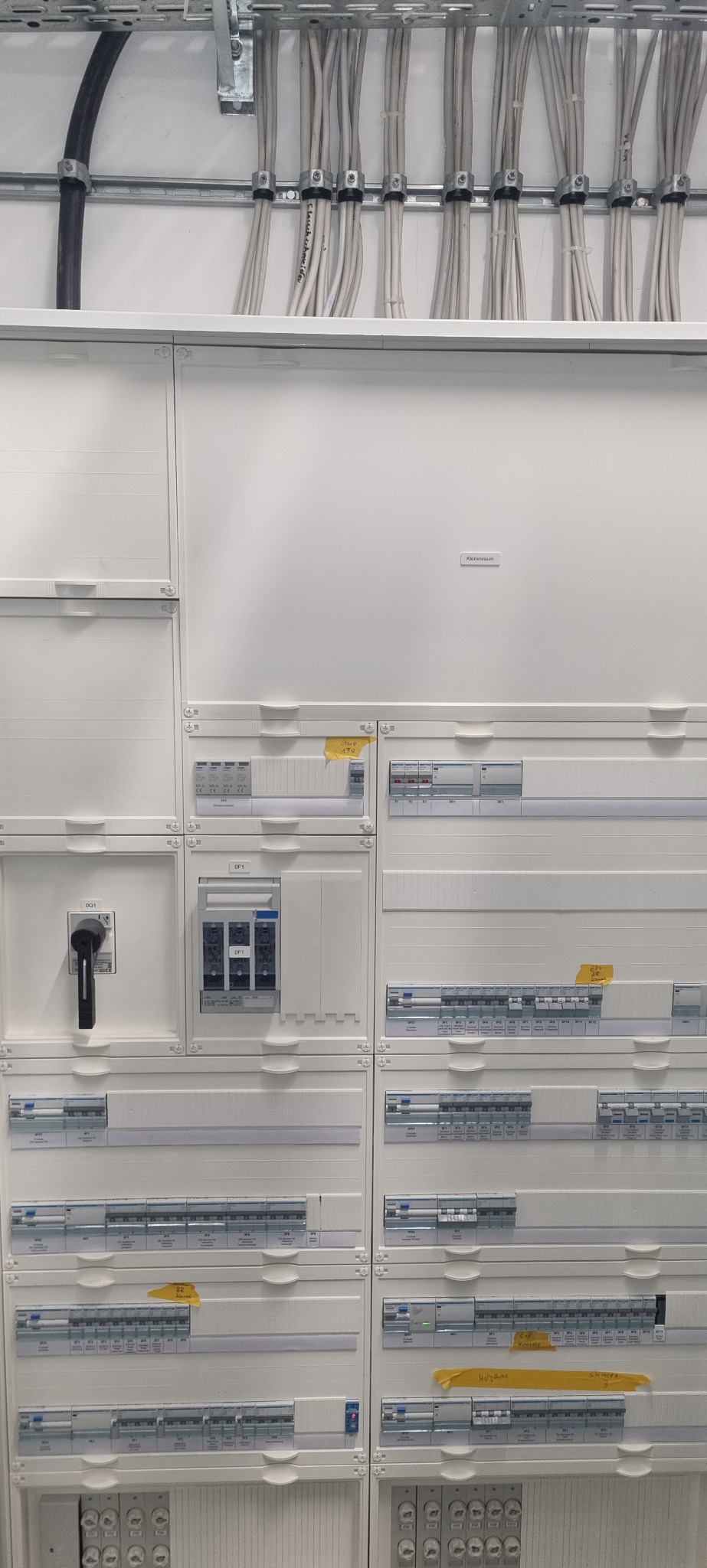 Rozdzielnica elektryczna z licznymi zabezpieczeniami, okablowaniem w korytkach i oznaczeniami na żółtych etykietach.