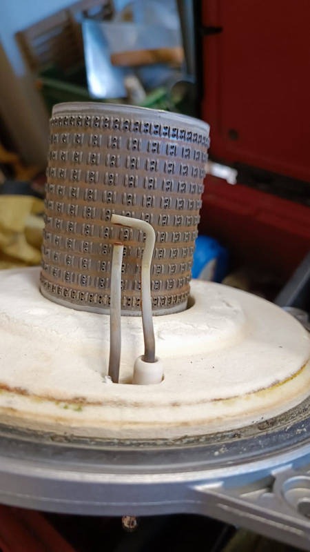 Ceramiczny cylinder z perforacją i dwoma metalowymi prętami, prawdopodobnie element pieca gazowego, widoczne ślady użytkowania i zabrudzenia.