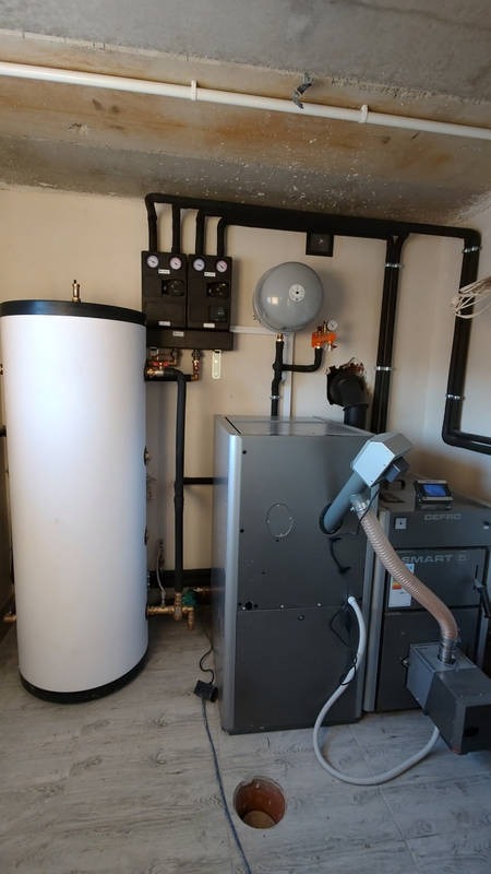 Kotłownia z piecem na pellet, zbiornikiem akumulacyjnym i czarnymi rurami instalacji grzewczej na ścianie, widoczna dziura w podłodze.