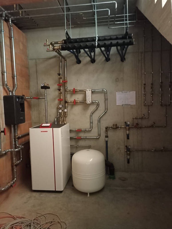 Zainstalowany, biały piec grzewczy marki Ochsner z czerwonym akcentem, obok biały zbiornik wyrównawczy. Widoczne liczne rury instalacji grzewczej z zaworami z czerwonymi pokrętłami, mocowane...