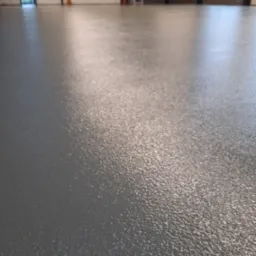 Gładka, szara posadzka w hali przemysłowej, widziana z niskiego kąta, z widoczną fakturą i refleksami światła, prowadząca do otwartych drzwi w tle.