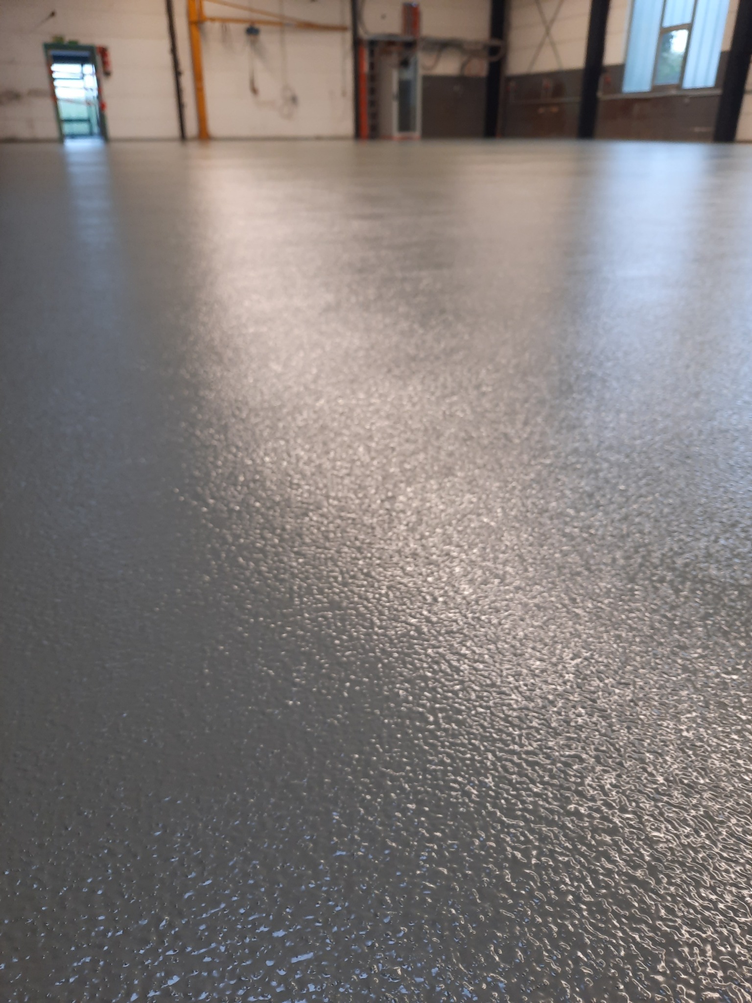 Gładka, szara posadzka w hali przemysłowej, widziana z niskiego kąta, z widoczną fakturą i refleksami światła, prowadząca do otwartych drzwi w tle.