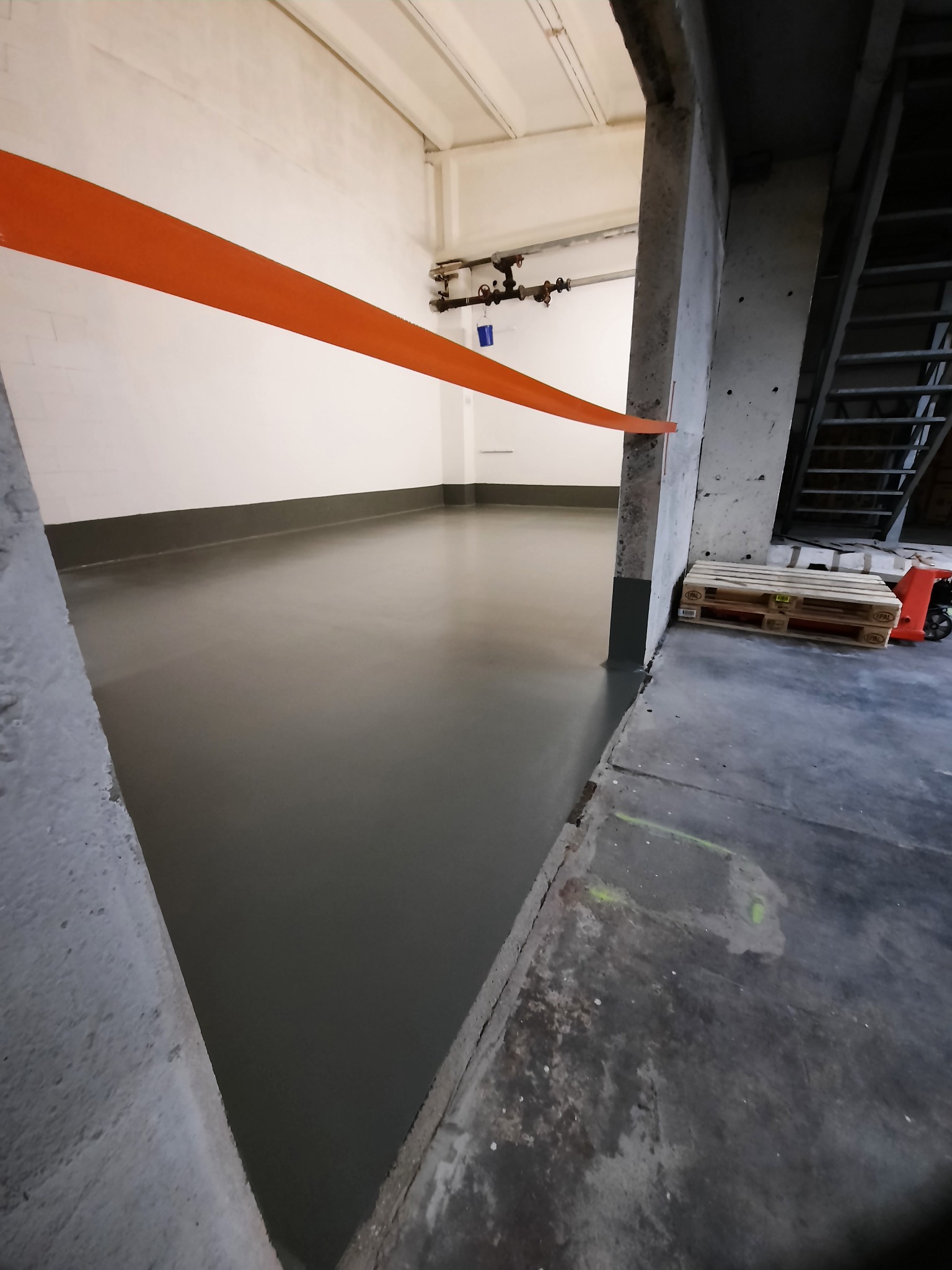 Świeżo wylana, szara posadzka w pomieszczeniu w Lęborku, zabezpieczona pomarańczową taśmą, widoczne palety i schody w tle.