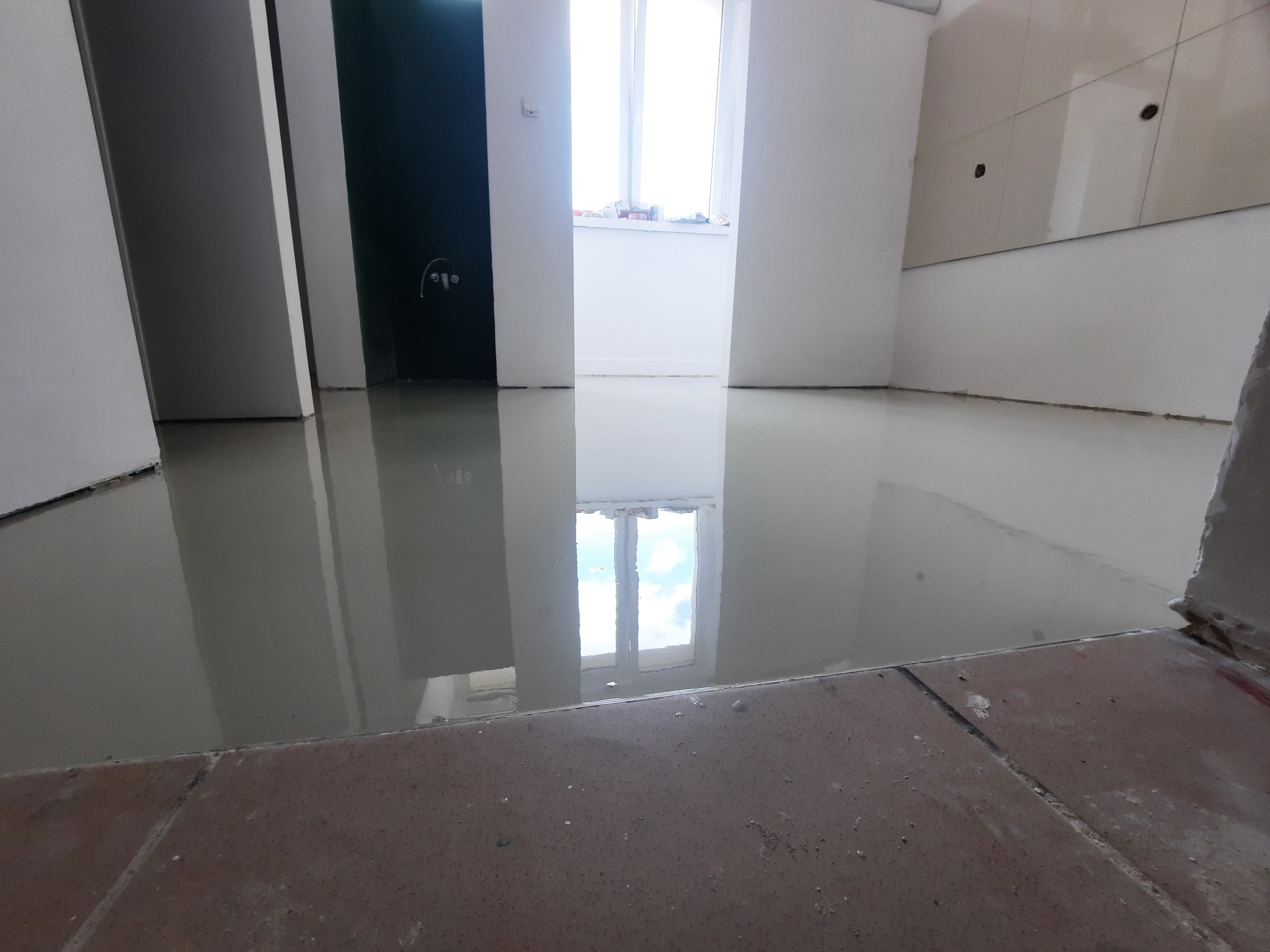 Świeżo wylana, samopoziomująca się wylewka w pomieszczeniu z widocznymi płytkami na podłodze w pierwszym planie i odbiciem okna w mokrej powierzchni.