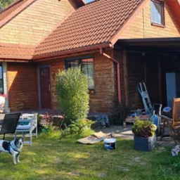 Malowanie drewnianego domu na zewnątrz: pracownik w trakcie renowacji elewacji, widoczne ślady starej farby, przygotowane narzędzia i farba, pies obserwuje prace na tle ogrodu.