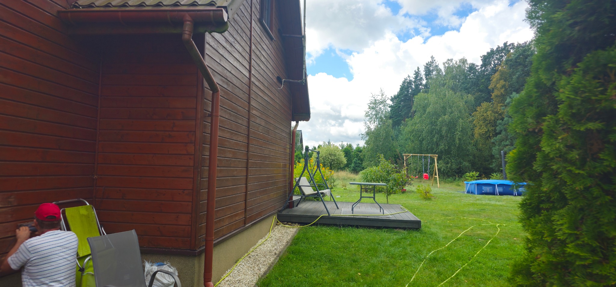 Mężczyzna w czerwonej czapce maluje drewnianą elewację domu na zewnątrz, widoczny fragment tarasu, trawnika i ogrodu z huśtawką i basenem w tle.