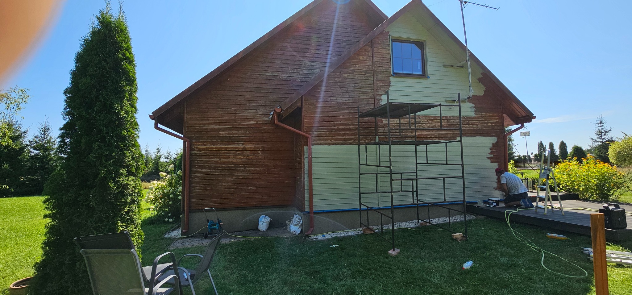 Malowanie elewacji drewnianego domu, widoczne rusztowanie i fragment pomalowanej ściany, pozostała część z surowego drewna, w tle zieleń ogrodu i błękit nieba.