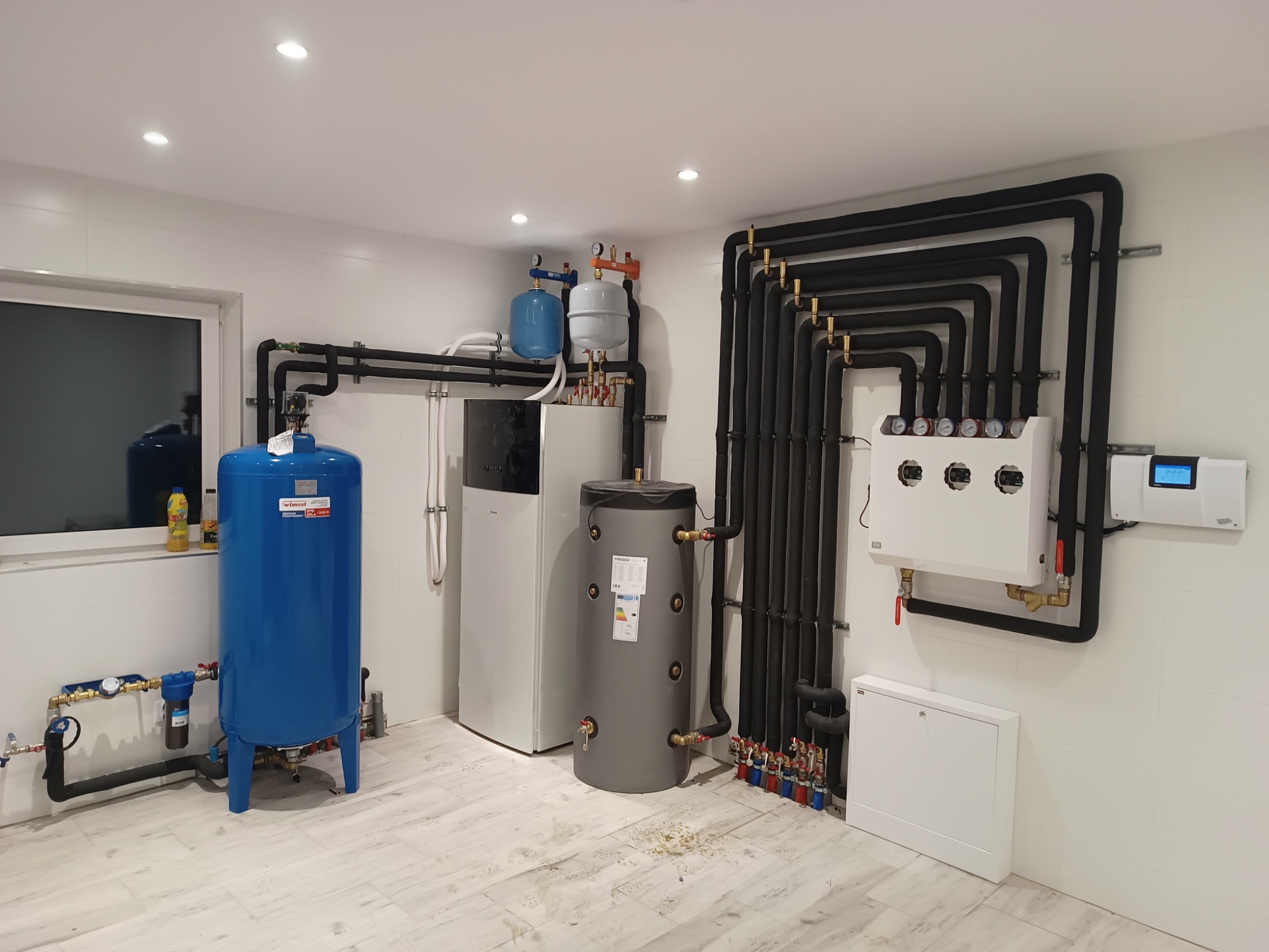 Kompletna instalacja grzewcza w kotłowni z szarym bojlerem, niebieskim zbiornikiem buforowym, pompą ciepła i rozbudowanym systemem rur w izolacji oraz aparaturą kontrolno-pomiarową na ścianie...