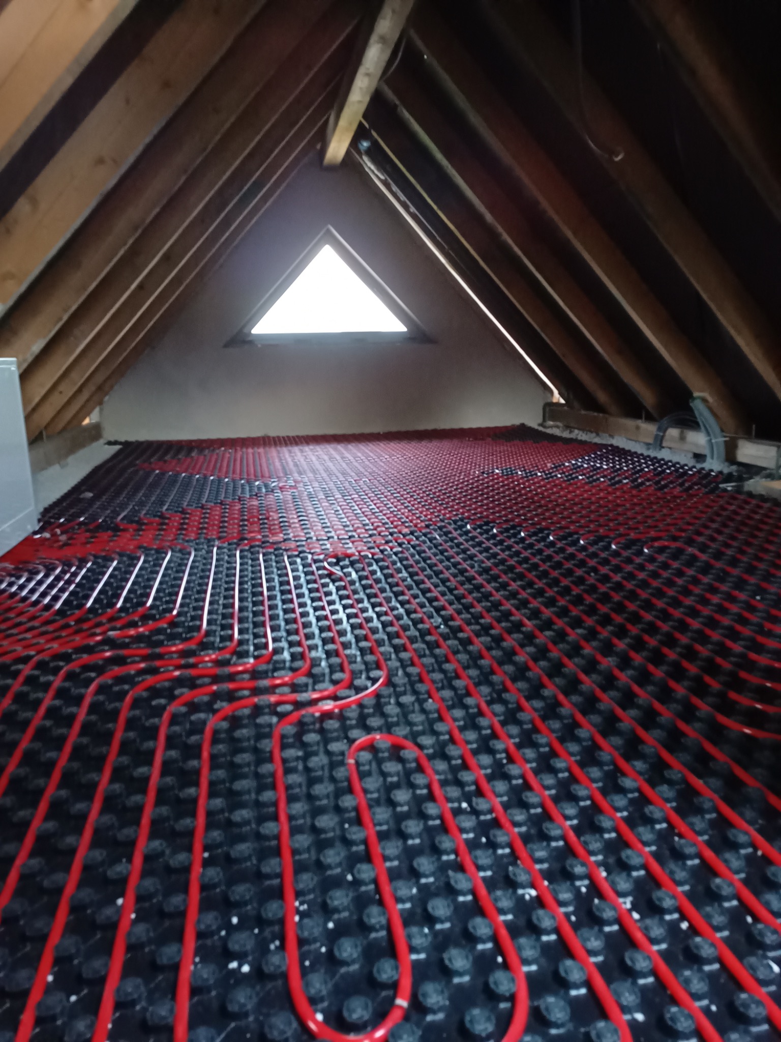 Instalacja ogrzewania podłogowego w trakcie montażu na poddaszu, widoczne czerwone rury ułożone na czarnej macie z wypustkami, drewniana konstrukcja dachu i okno trójkątne w tle.
