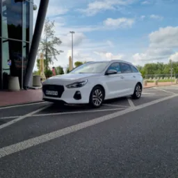 Białe kombi zaparkowane przed nowoczesnym budynkiem z przeszkloną fasadą, widoczne linie parkingu i fragment błękitnego nieba z chmurami.