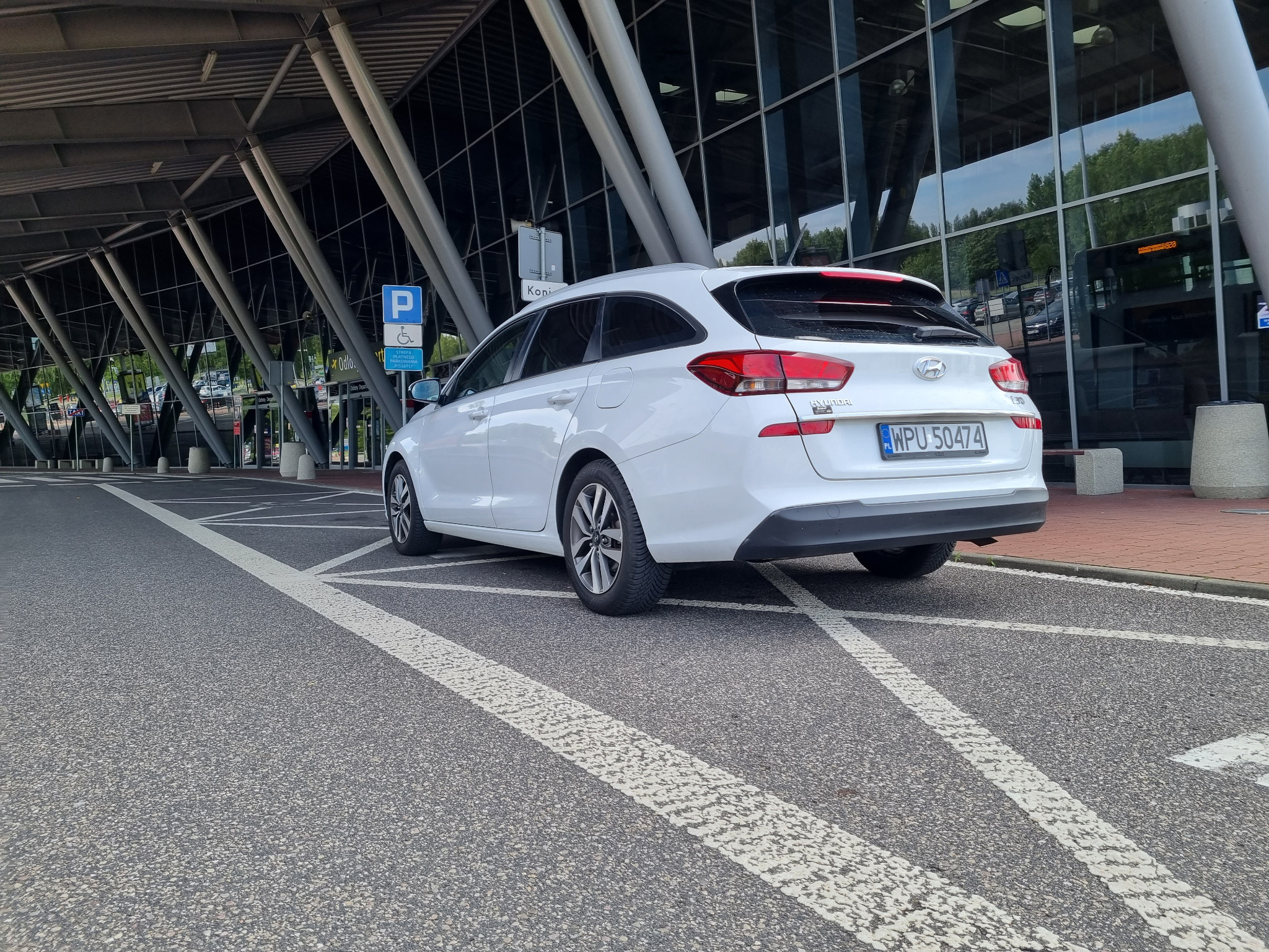 Biały samochód kombi Hyundai i30 zaparkowany na parkingu lotniska, widok z tyłu, tablica rejestracyjna WPU 50474, znak parkingowy dla niepełnosprawnych w tle.
