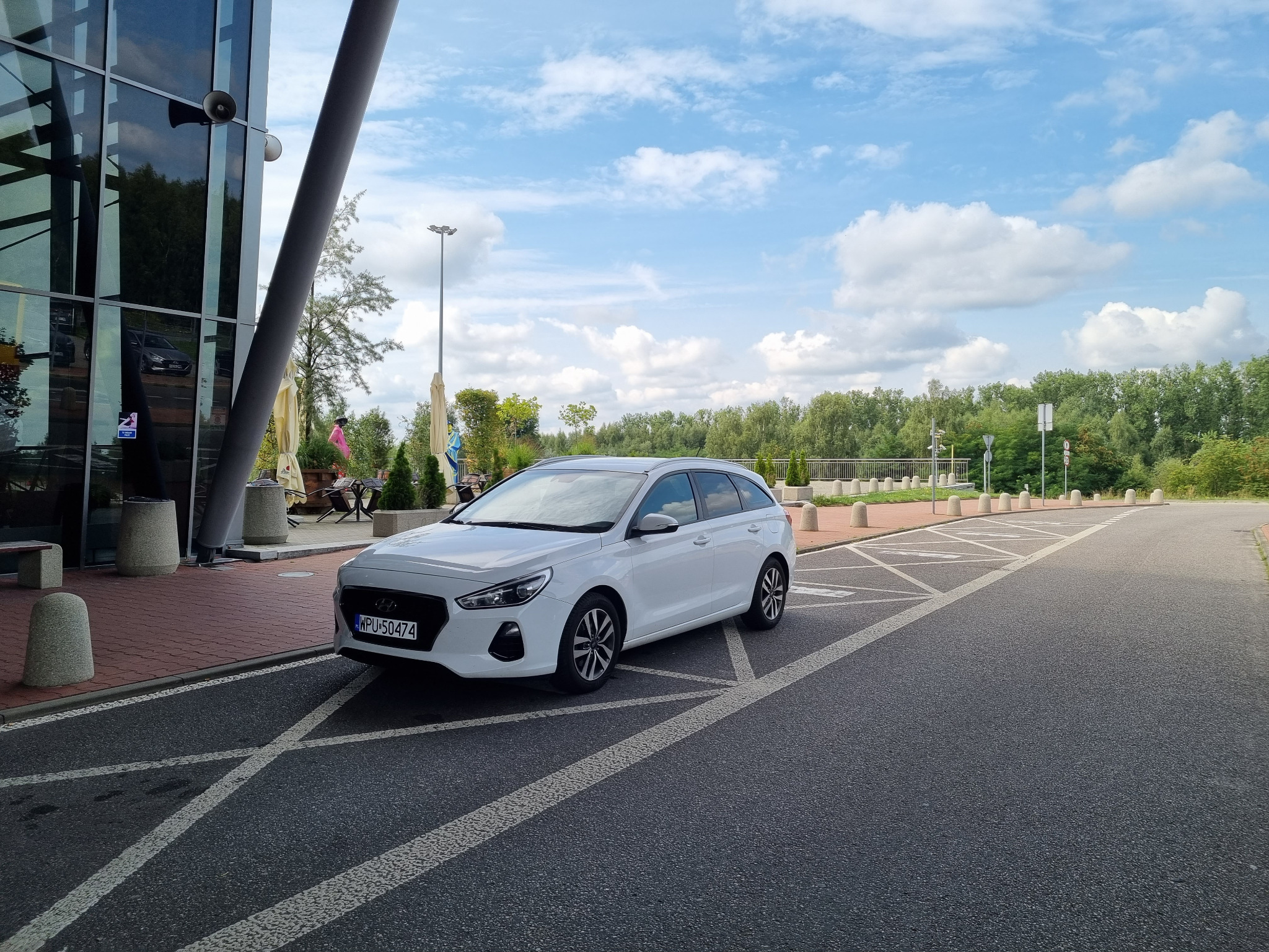 Biały samochód kombi zaparkowany na parkingu z namalowanymi liniami, na tle nowoczesnego budynku z elementami szkła i metalu oraz drzew