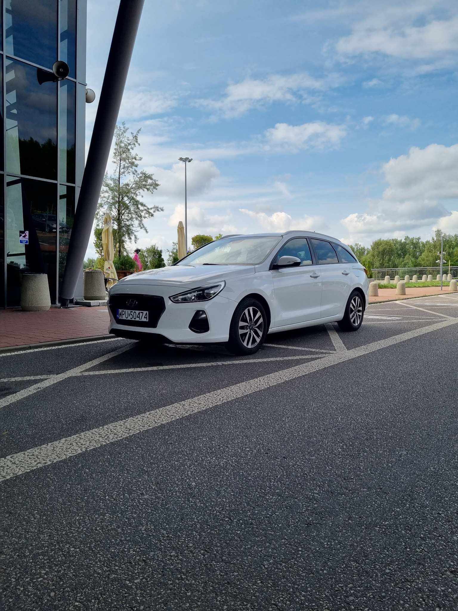 Białe kombi zaparkowane przed nowoczesnym budynkiem z przeszkloną fasadą, widoczne linie parkingu i fragment błękitnego nieba z chmurami.