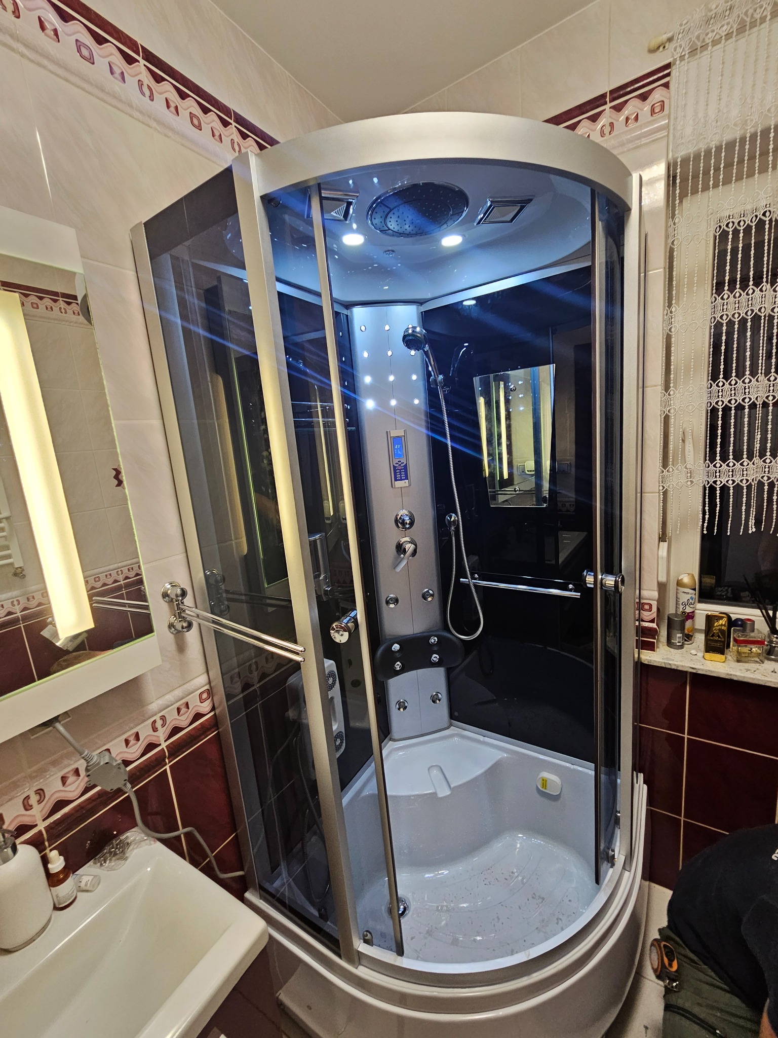 Nowoczesna kabina prysznicowa z hydromasażem, oświetleniem LED i panelem sterowania, zamontowana w łazience z brązowo-białymi kafelkami, widok z przodu na otwarte drzwi kabiny, z boku umywalka...