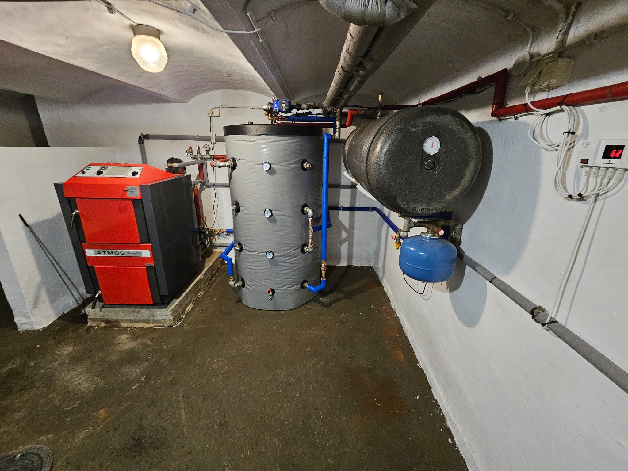 Kotłownia z kotłem na paliwo stałe Atmos, zbiornikiem akumulacyjnym i zasobnikiem ciepłej wody użytkowej, rury w kolorze czerwonym i niebieskim, widoczne manometry i sterownik temperatury.