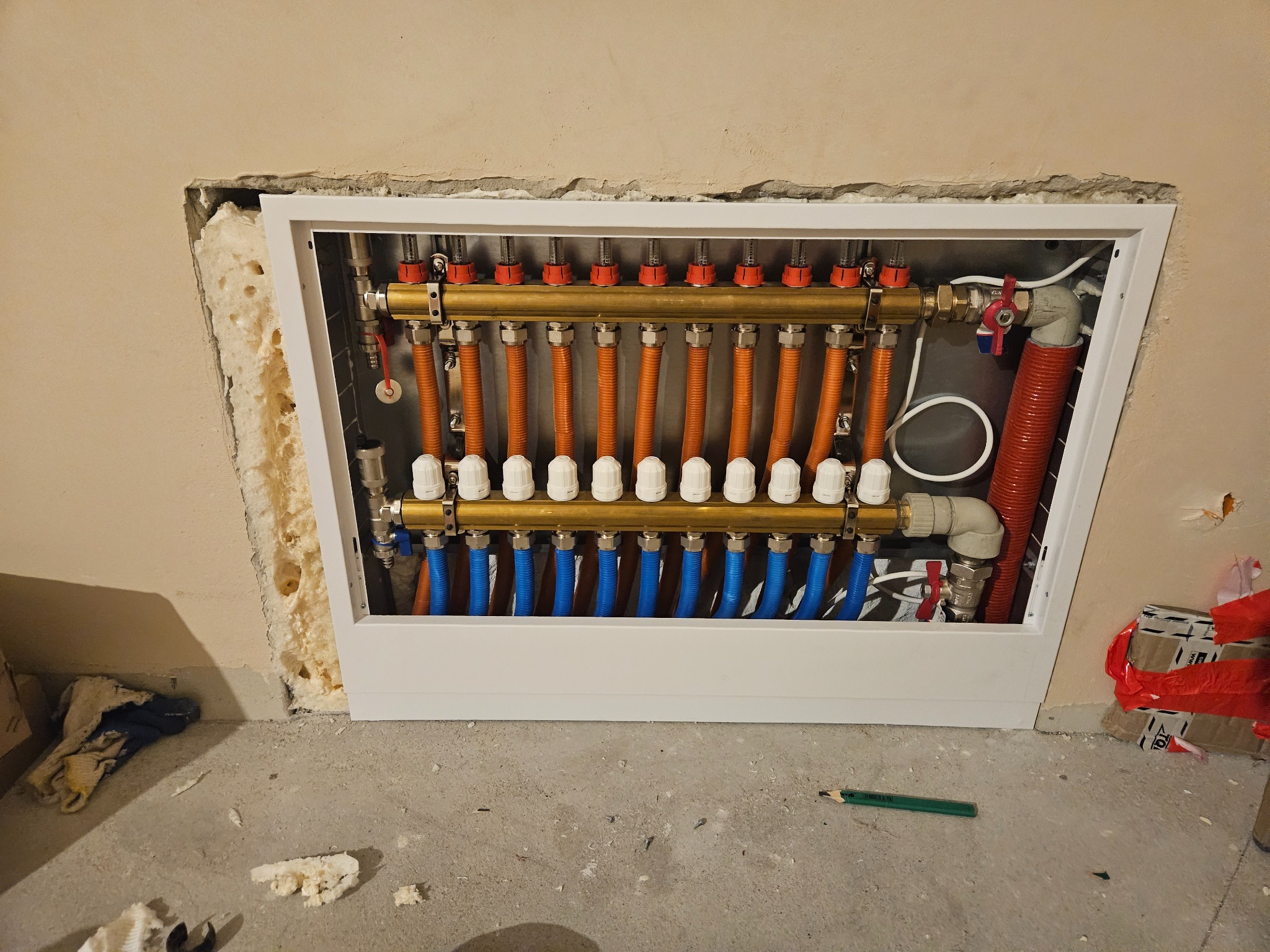 Rozdzielacz ogrzewania podłogowego z rurami w kolorach pomarańczowym i niebieskim, zamontowany w ścianie z widoczną izolacją, na podłodze leży ołówek stolarski.