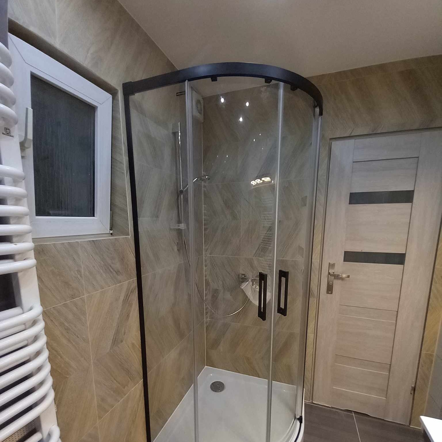 Kabina prysznicowa z czarną ramą w narożniku łazienki z beżowymi płytkami, widoczny grzejnik drabinkowy i drzwi z poziomymi panelami.