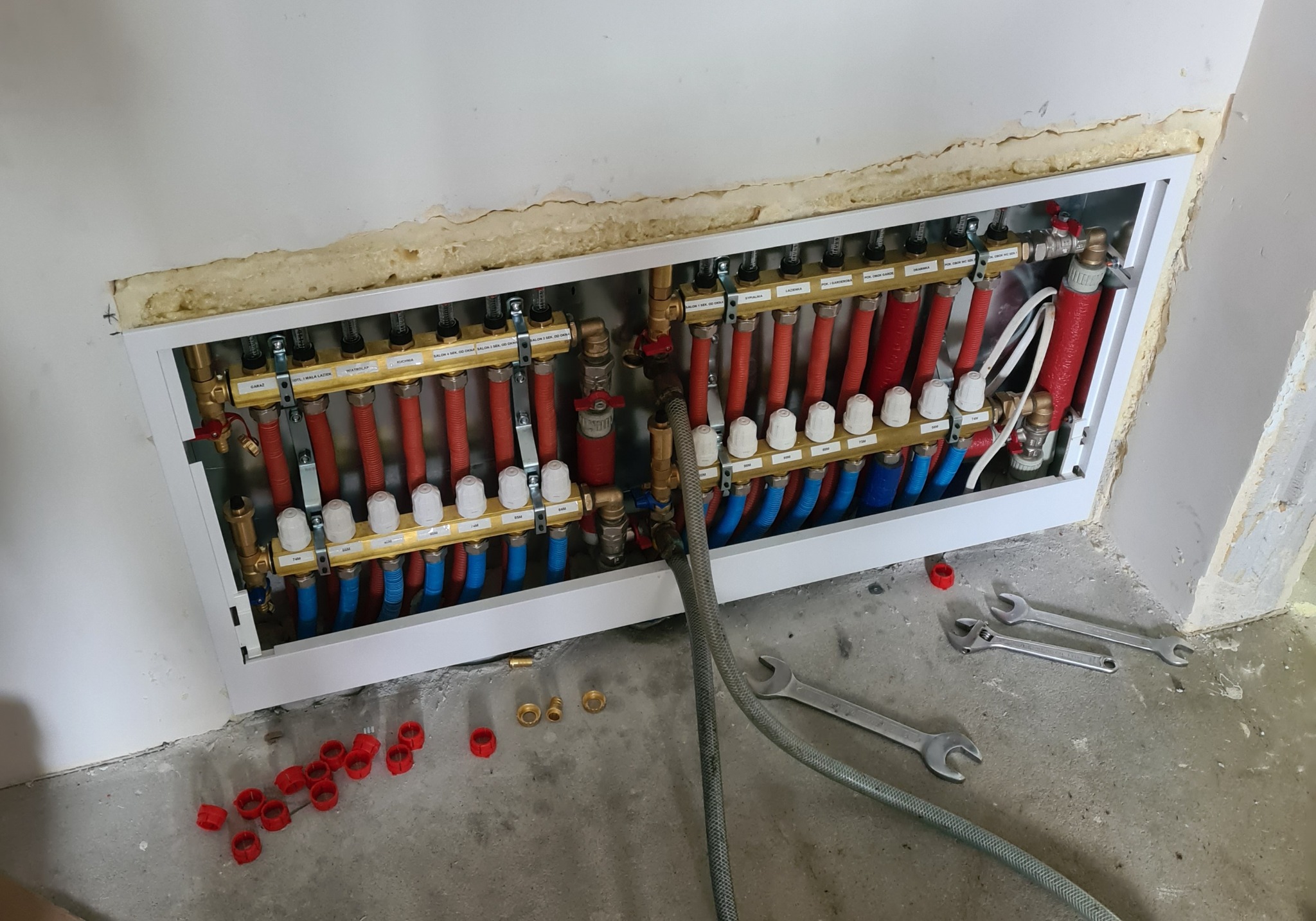 Rozdzielacz ogrzewania podłogowego w obudowie, z widocznymi rurami czerwonymi i niebieskimi, manometry, zawory, oraz klucze i złączki na podłodze w trakcie montażu.