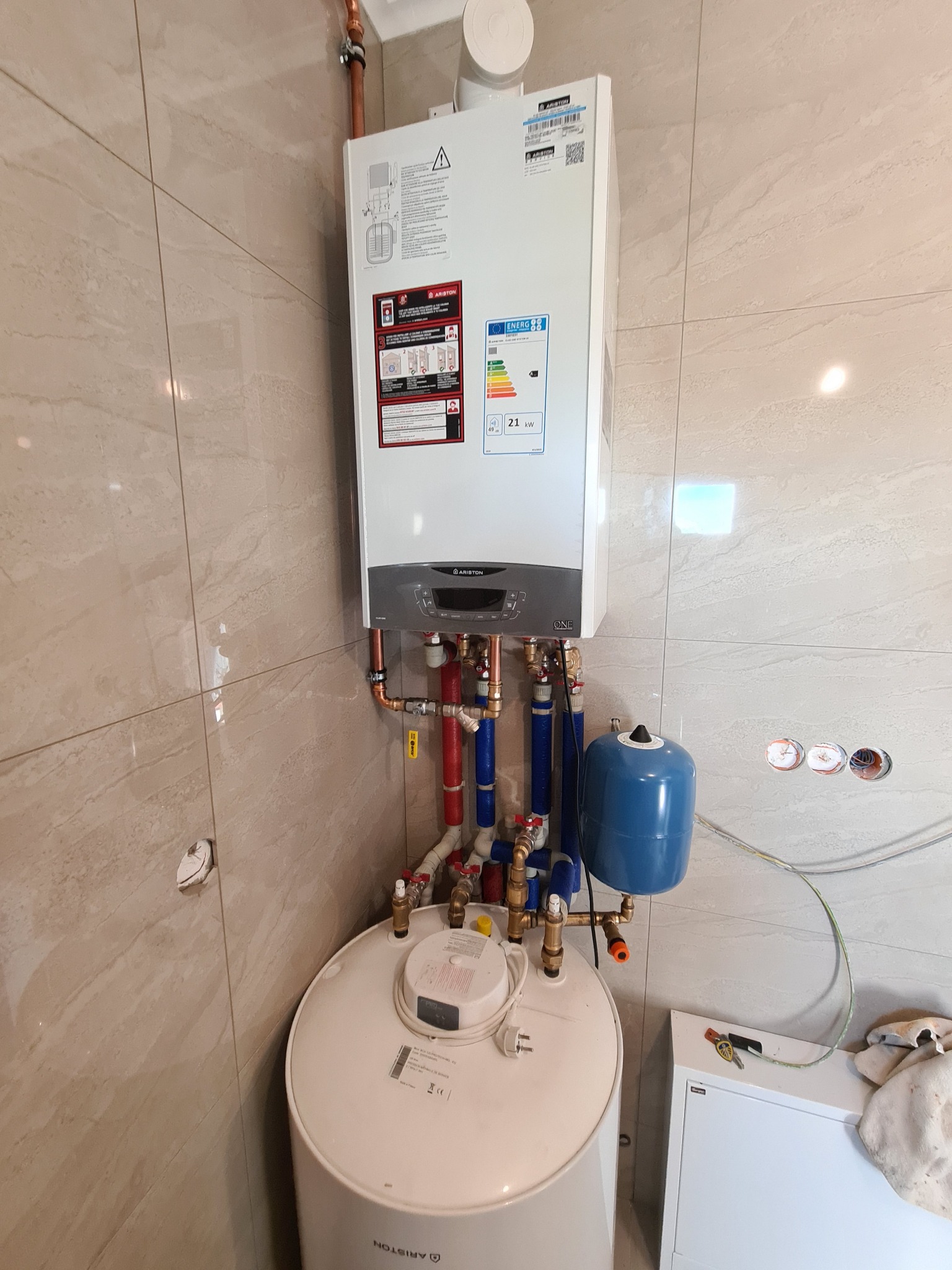 Kocioł gazowy Ariston ONE wiszący na ścianie z beżowych płytek, podłączony do zasobnika ciepłej wody użytkowej i instalacji, z widocznymi rurami miedzianymi, niebieskimi i czerwonymi,...