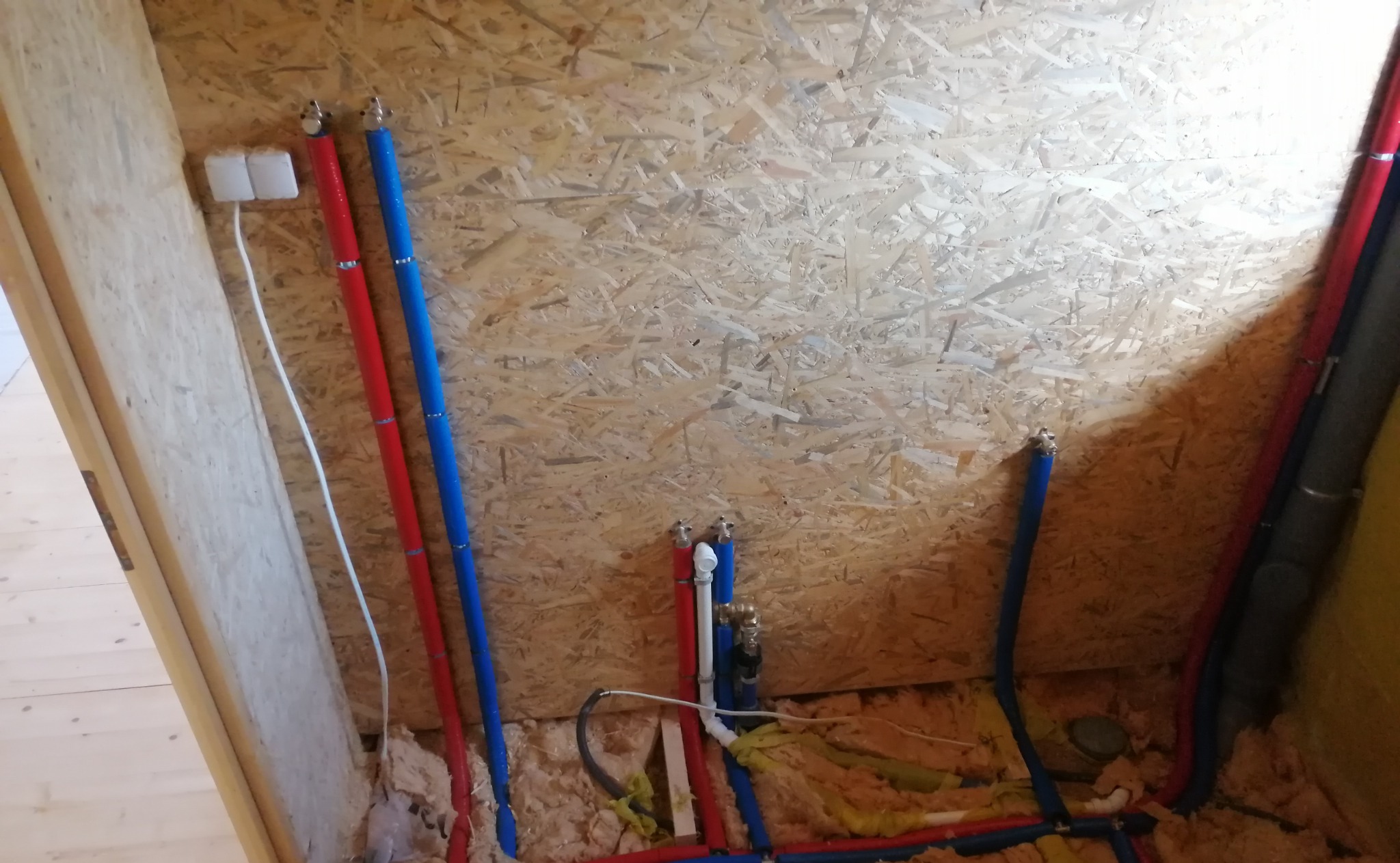 Wewnętrzna instalacja wodno-kanalizacyjna w trakcie montażu, widoczne rury PEX w kolorze niebieskim i czerwonym, izolacja z wełny mineralnej oraz ściana z płyty OSB.