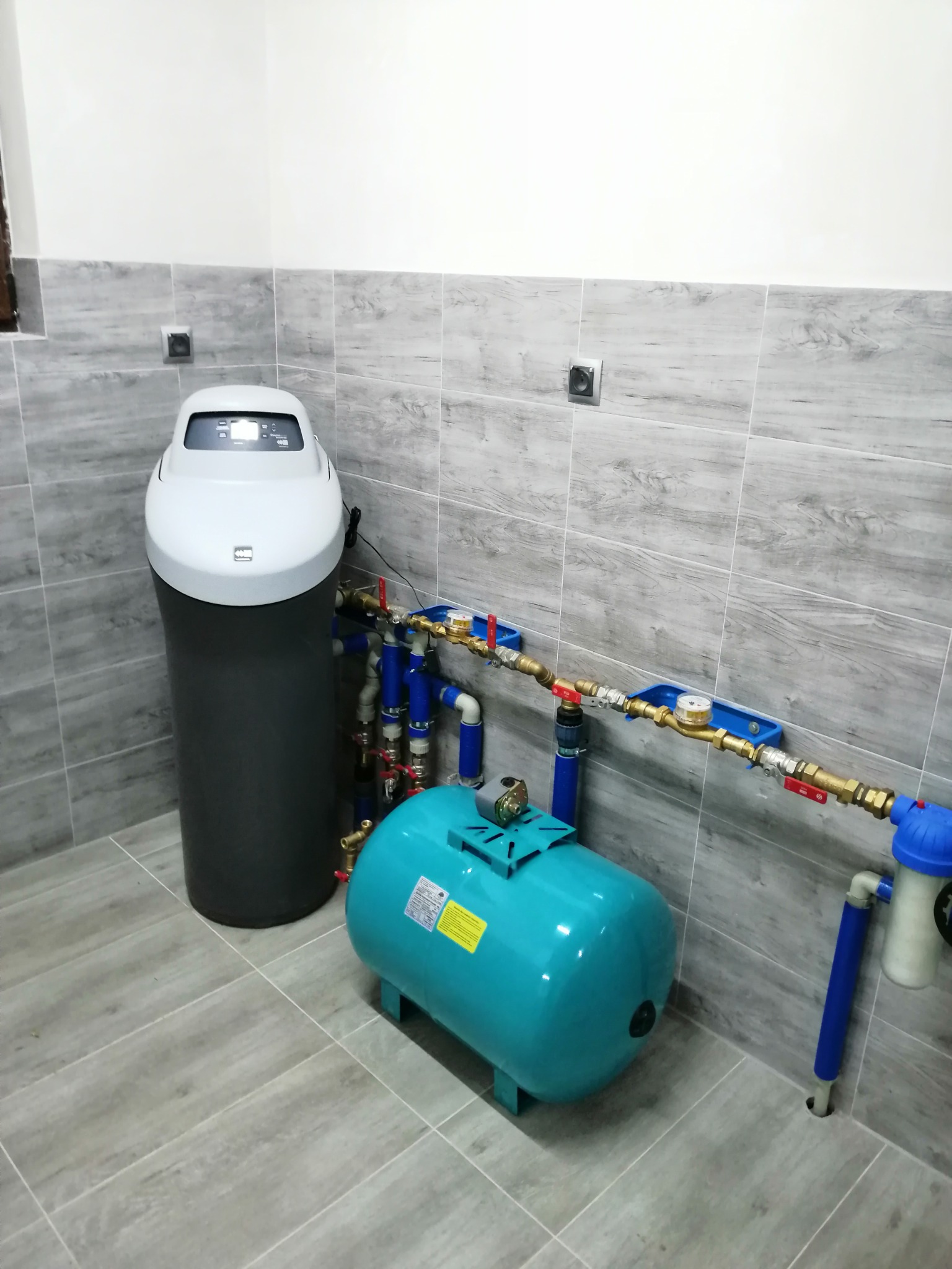 Zestaw do uzdatniania wody: duży filtr kolumnowy i zbiornik ciśnieniowy w turkusowym kolorze, połączone rurami w pomieszczeniu z szarymi płytkami imitującymi drewno.