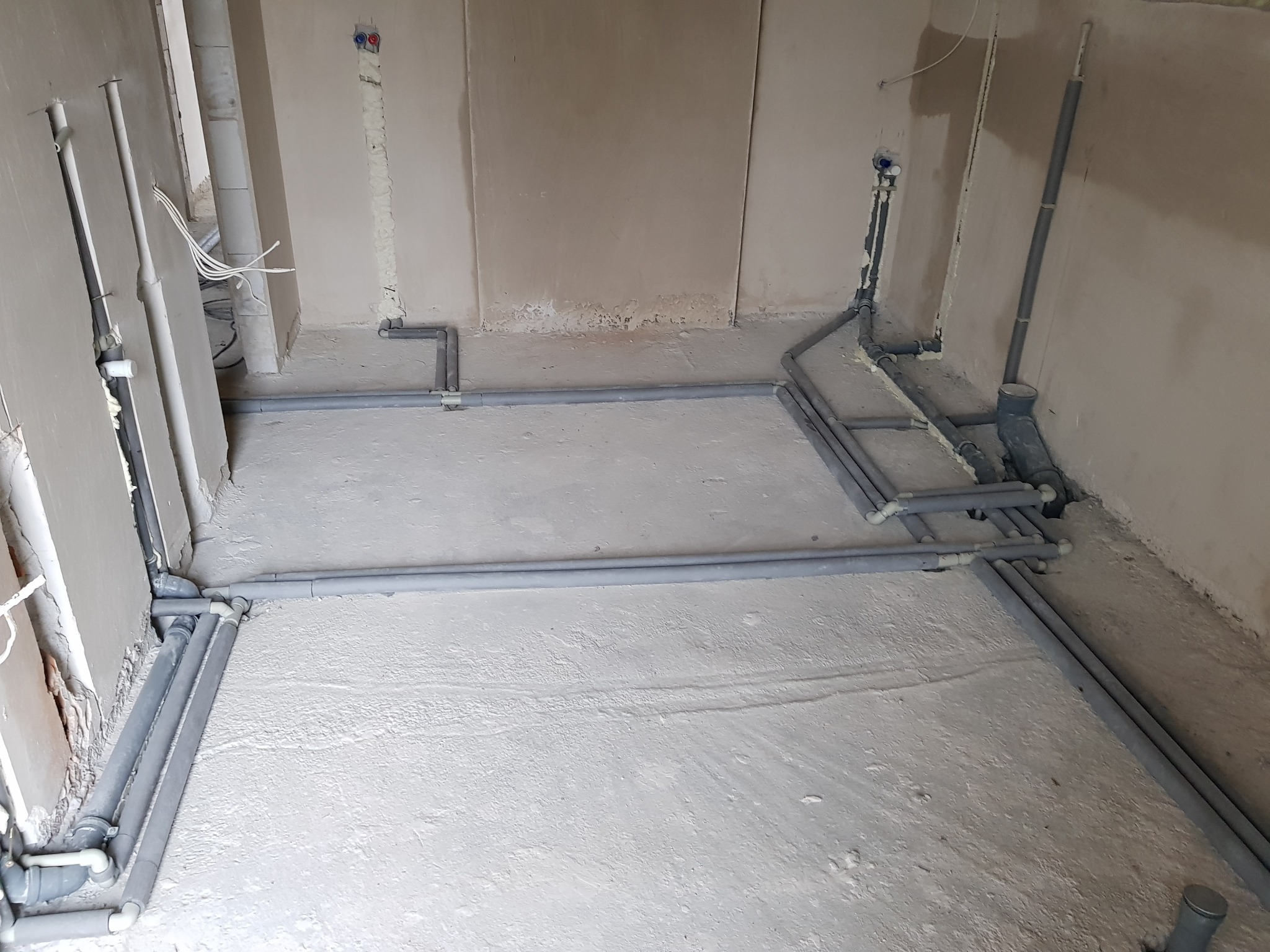 Szare rury kanalizacyjne ułożone na surowej posadzce w trakcie montażu instalacji w surowym stanie. Widoczne połączenia i pianka montażowa przy ścianach.