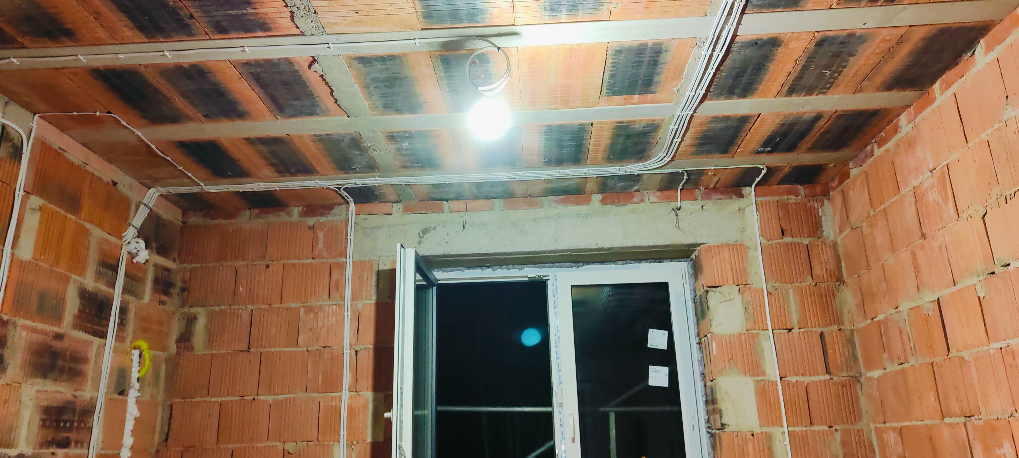 Białe przewody elektryczne prowadzone po ceglanej ścianie i suficie w surowym stanie, tymczasowe oświetlenie żarówką, okno z widokiem na noc.