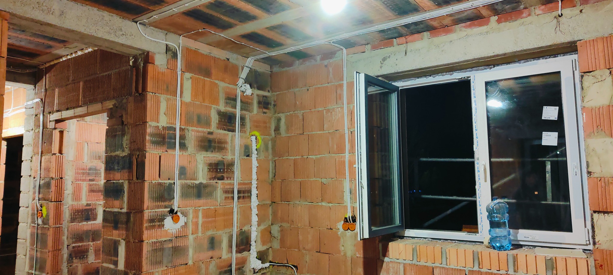 Wnętrze w trakcie remontu: widoczne przewody elektryczne prowadzone po surowych ścianach z cegły, częściowo otynkowane, z zamontowanym oknem PCV i butelką wody na parapecie.