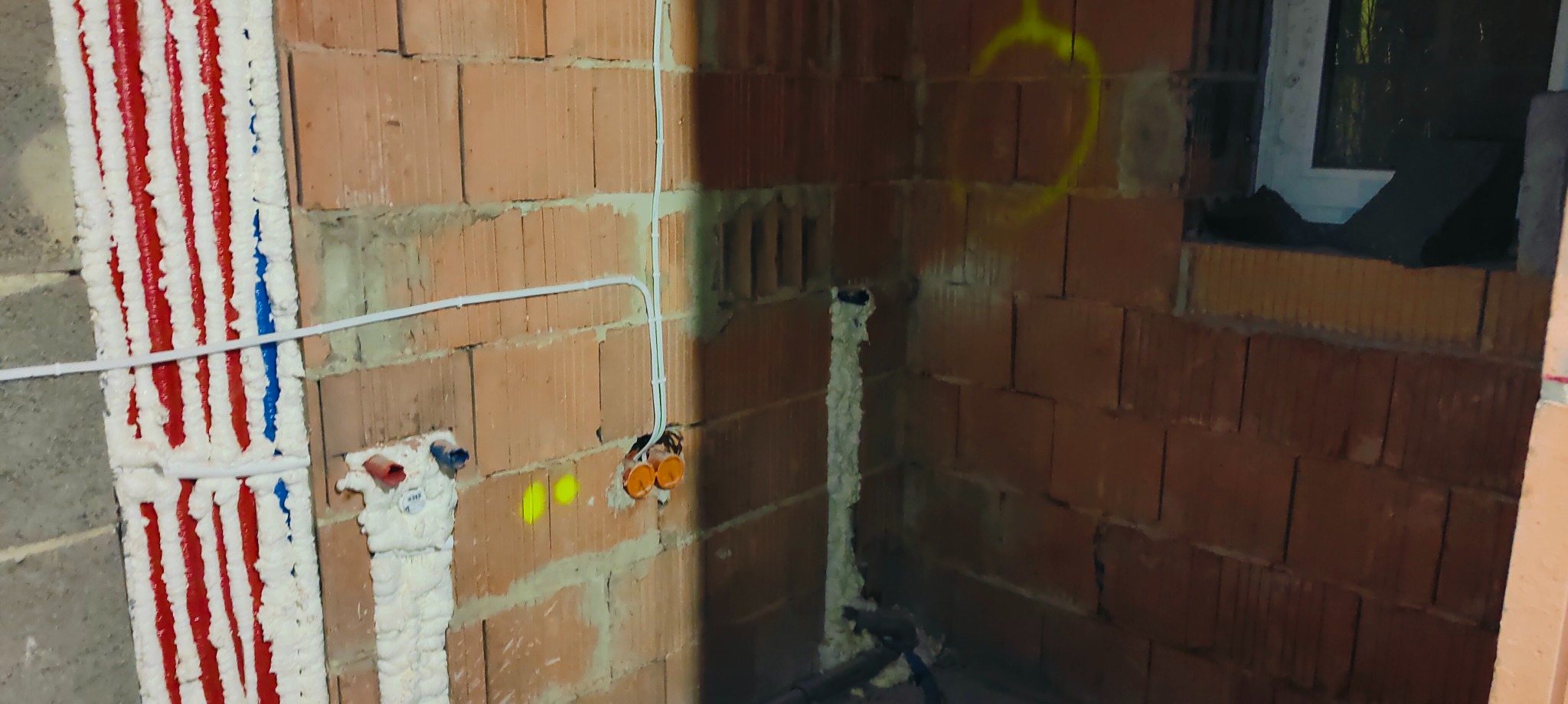 Surowy stan wnętrza z ceglanymi ścianami, widoczne rury instalacji grzewczej zabezpieczone pianką montażową, przewody elektryczne w peszlach oraz okno w trakcie montażu.
